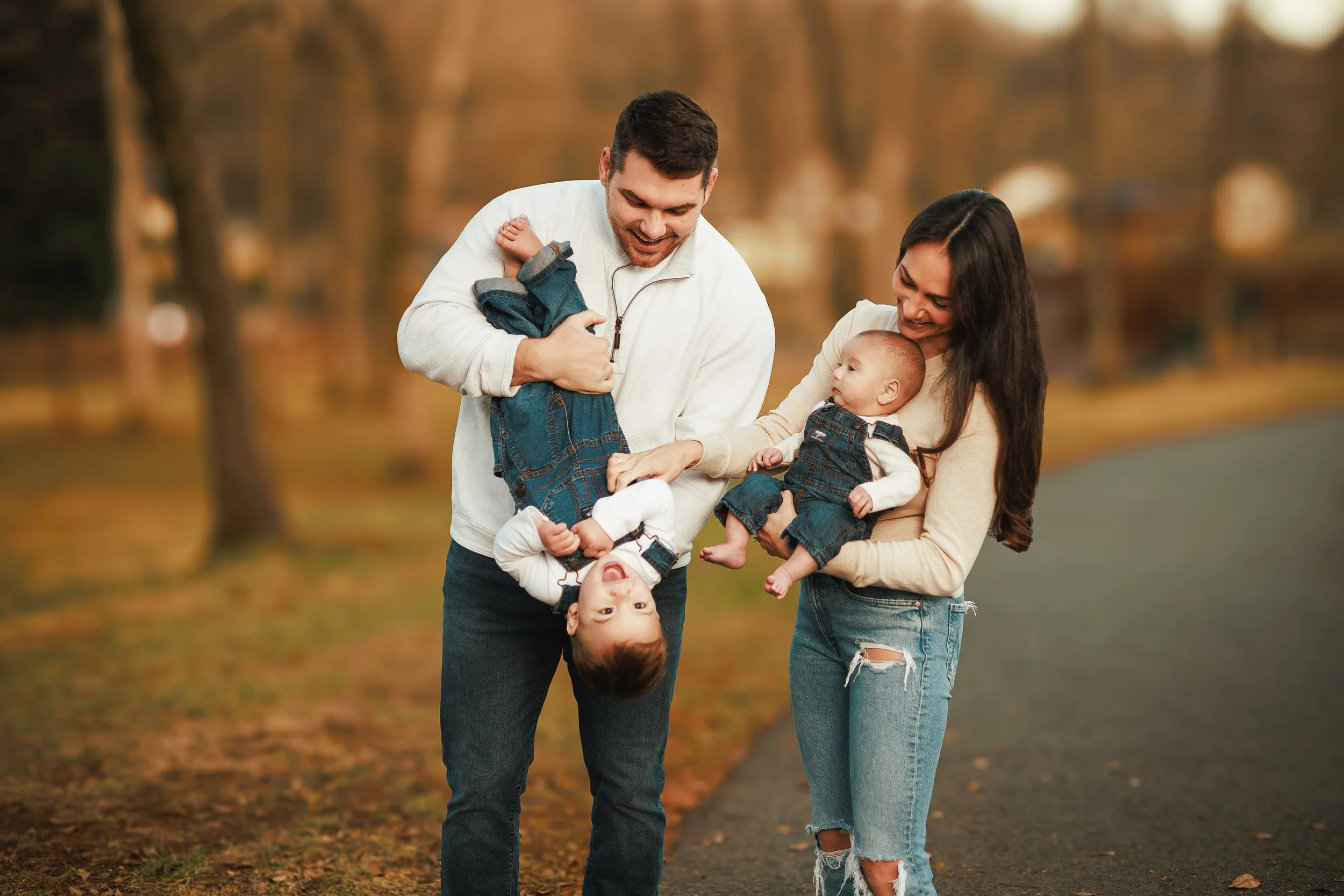 A family of four, including a father, mother, and two young children, enjoying a walk in a park during autumn. The father is holding a young boy upside down, and the mother is holding a baby girl, all smiling and appearing happy.