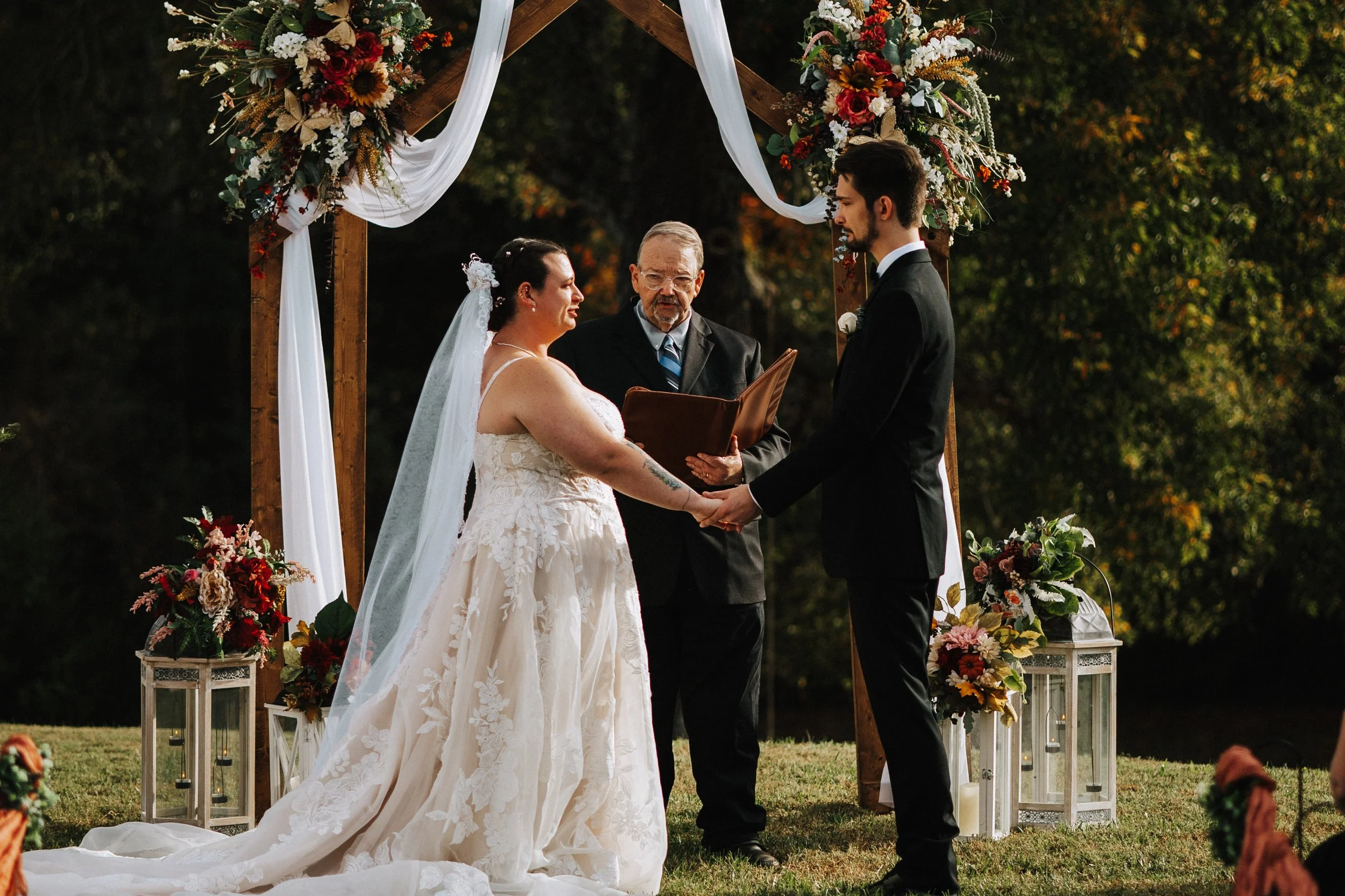 A couple gets married outdoors under a wooden arch decorated with flowers and white fabric, with a man officiating the ceremony. The bride is in a white lace gown with a veil, and the groom is in a black suit. There are lamps on the ground in front o