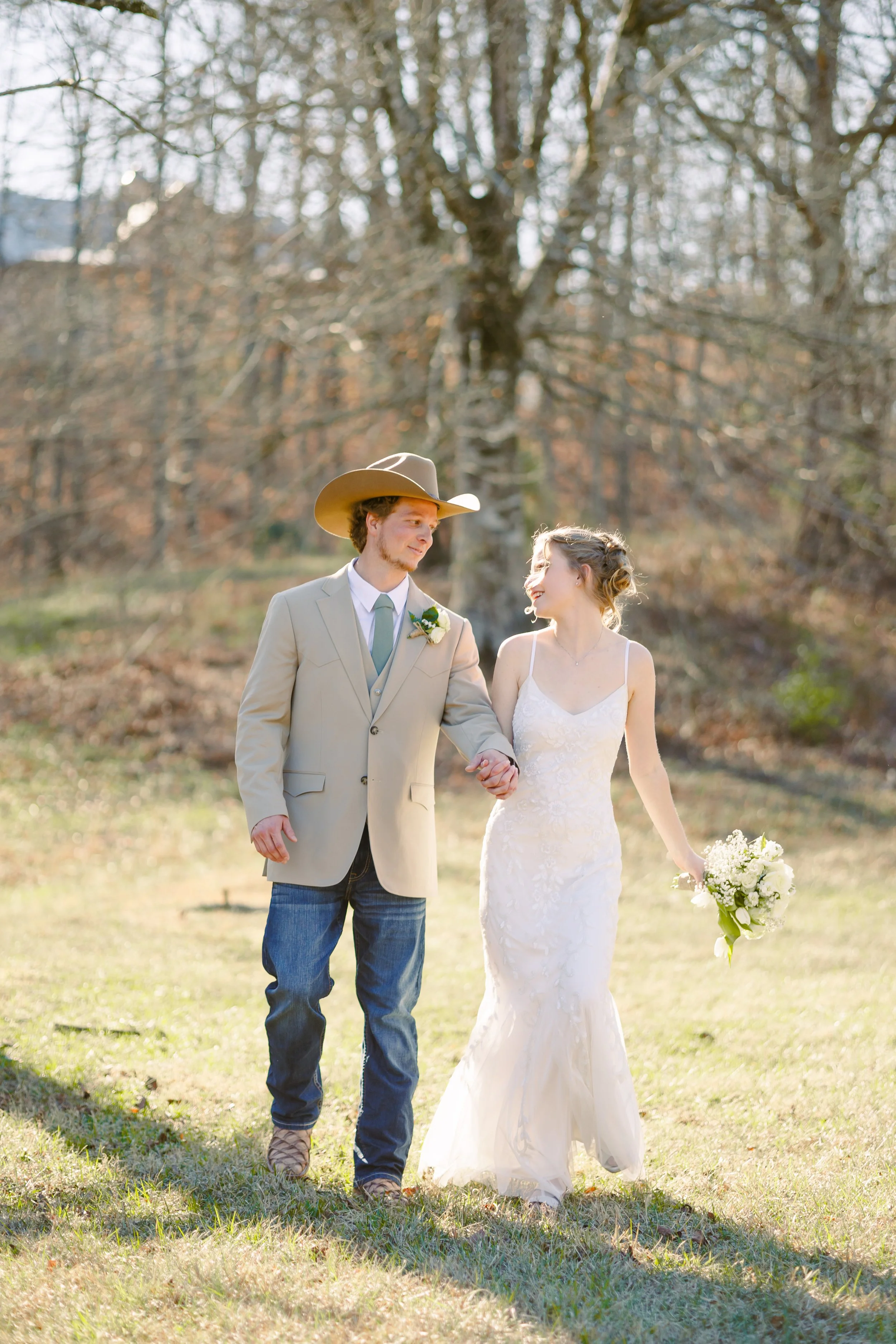A bride and groom holding hands and walking outdoors in a wooded area during daylight, with the bride wearing a white lace wedding dress and holding a bouquet, and the groom dressed in a beige suit jacket, blue jeans, and a cowboy hat.
