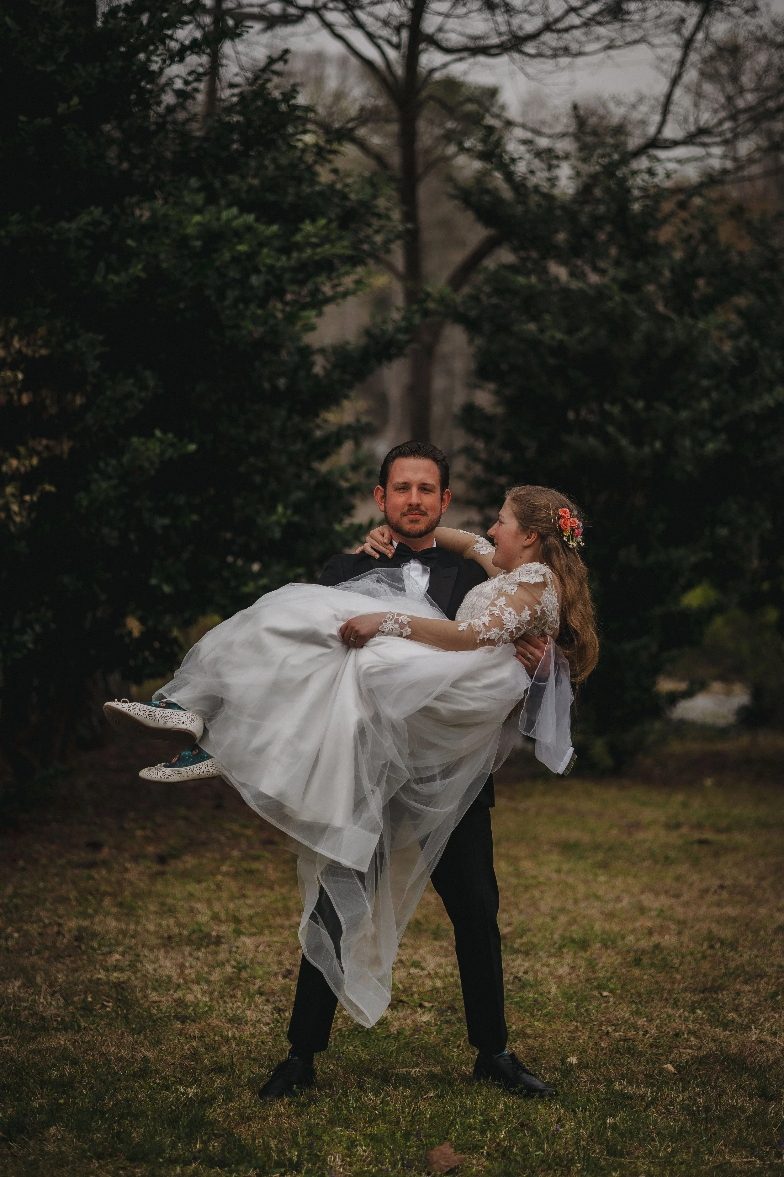 A man in a tuxedo holding a woman in a wedding dress outdoors surrounded by trees.