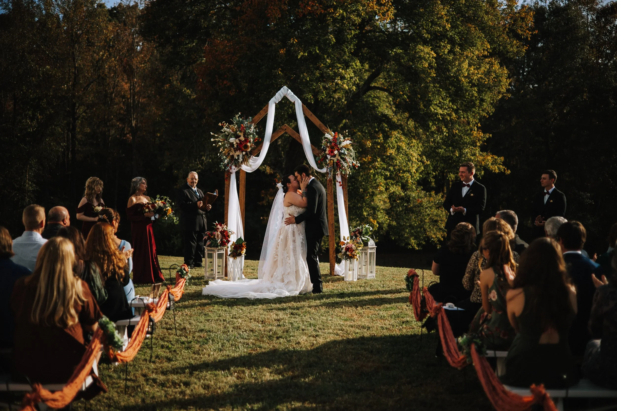 A couple in wedding attire kissing under a decorated wooden arch at an outdoor wedding ceremony surrounded by guests and lush trees.