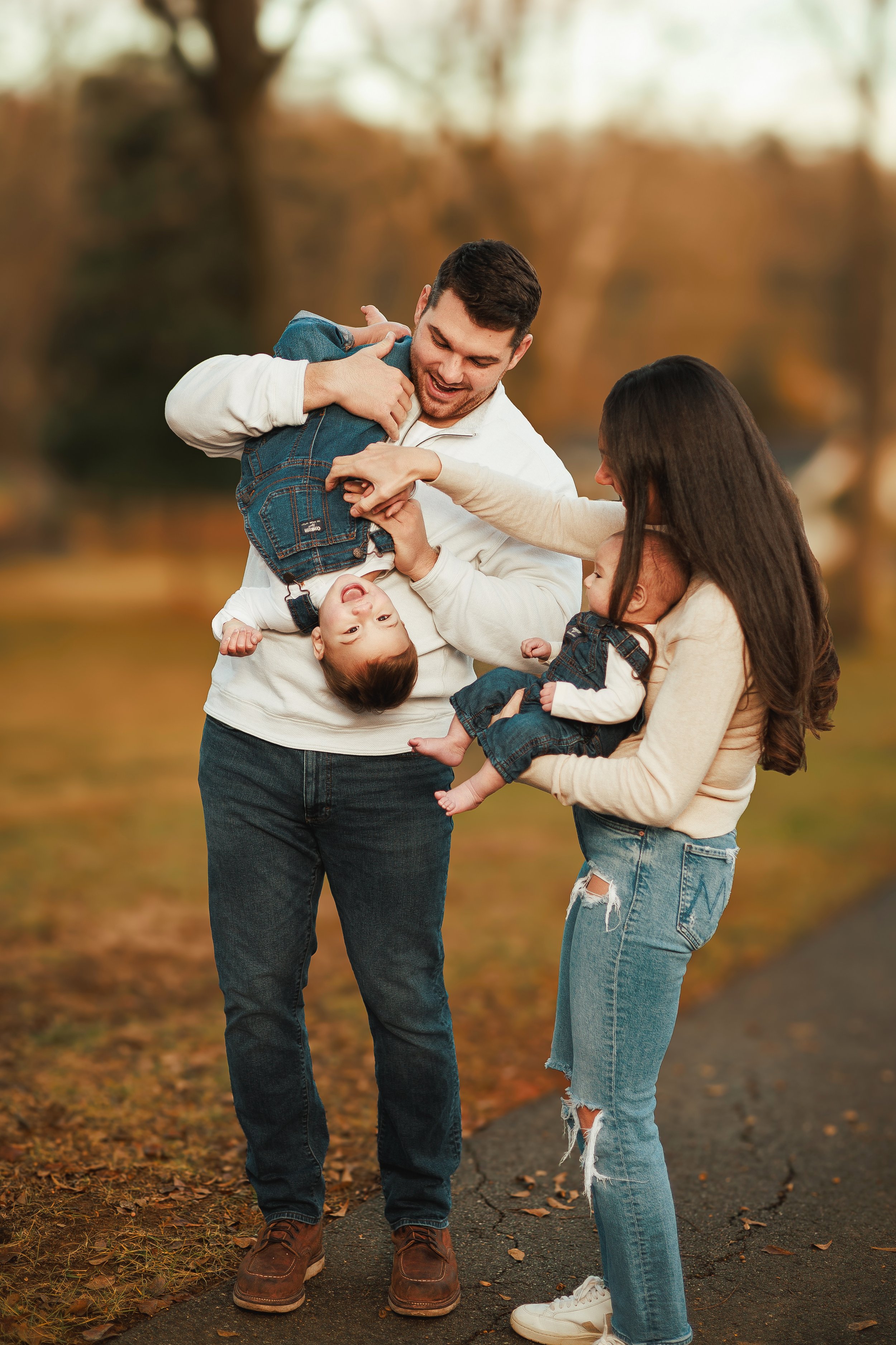 Family of four - father, mother, and two children - playing outdoors on a fall day, with trees and leaves around.