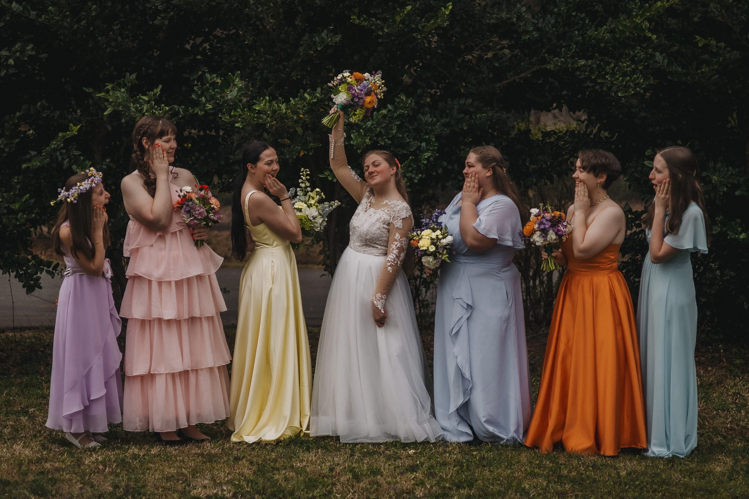 A bride and six women stand outdoors in front of green foliage, wearing colorful dresses. The bride holds a bouquet and is smiling, surrounded by women who are reacting with surprise or admiration, some touching their faces or holding flowers.