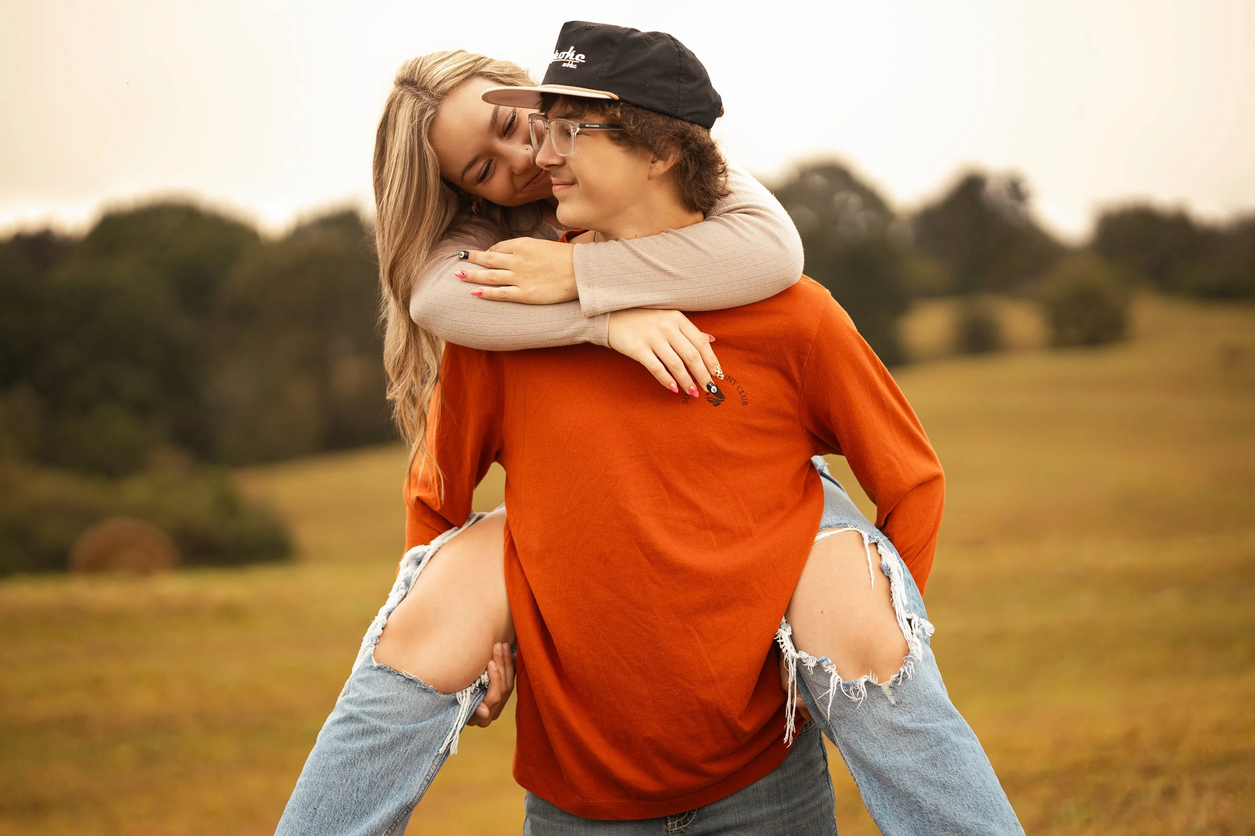 A woman giving a piggyback ride to a man outdoors during sunset or late afternoon with open field and trees in the background.