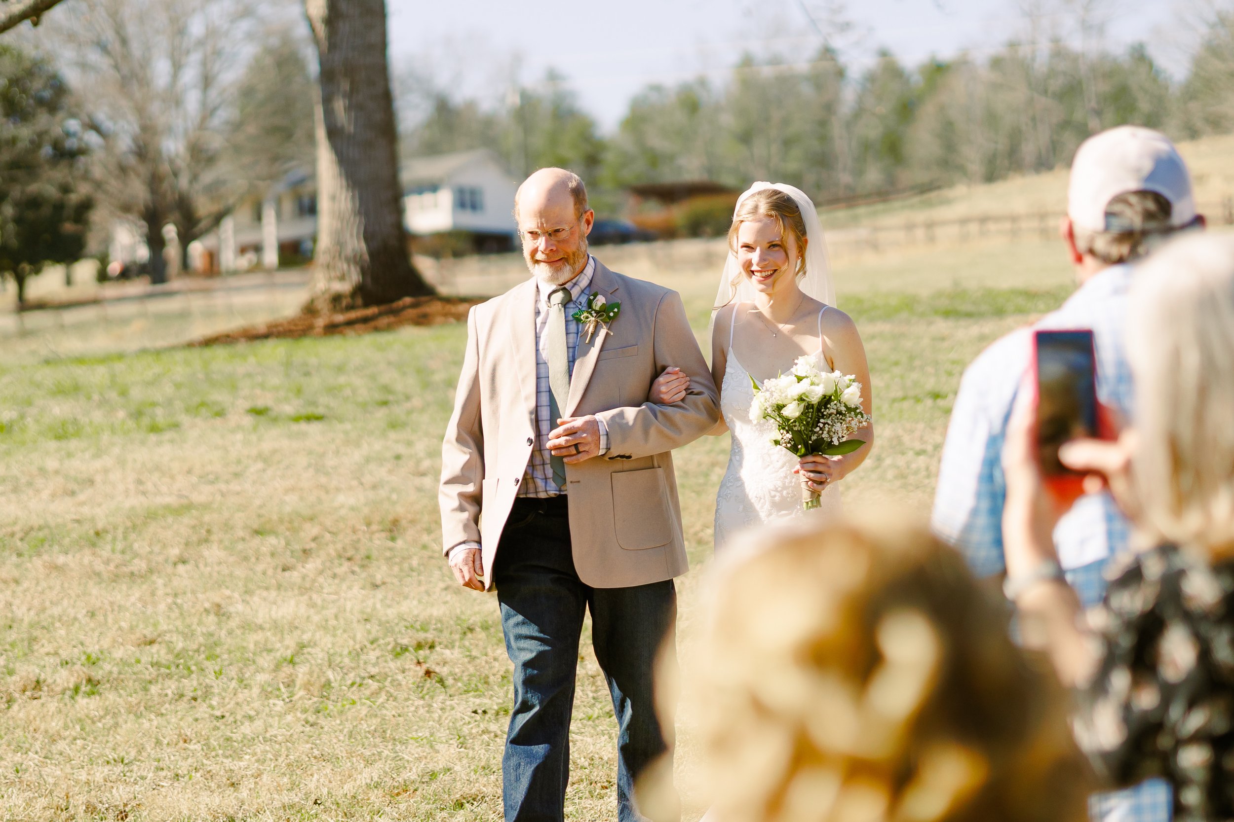 A bride in a white dress and holding a bouquet walks down the aisle with a man in a beige jacket and glasses, during an outdoor wedding ceremony. Guests are seen taking photos in the foreground, and there are houses and large trees in the background.