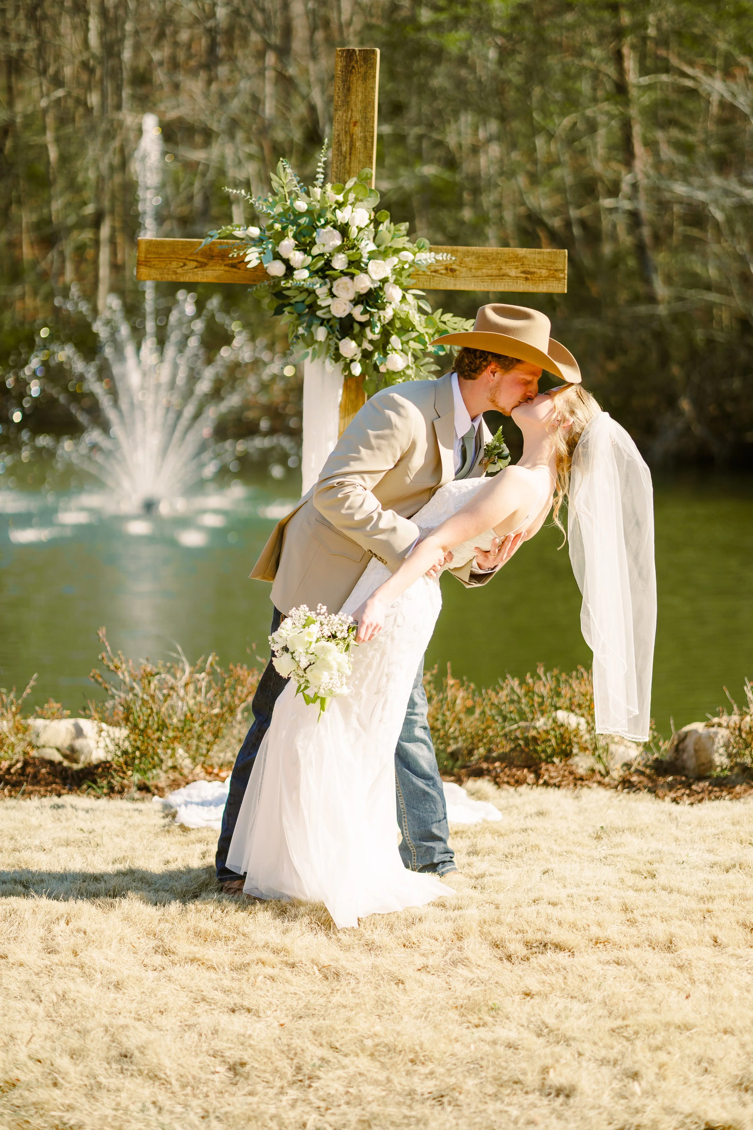 A newlywed couple sharing a kiss outdoors, with the groom dressed in a cowboy hat and beige suit, and the bride wearing a white wedding dress and veil, with a wooden cross decorated with white roses and greenery in the background.