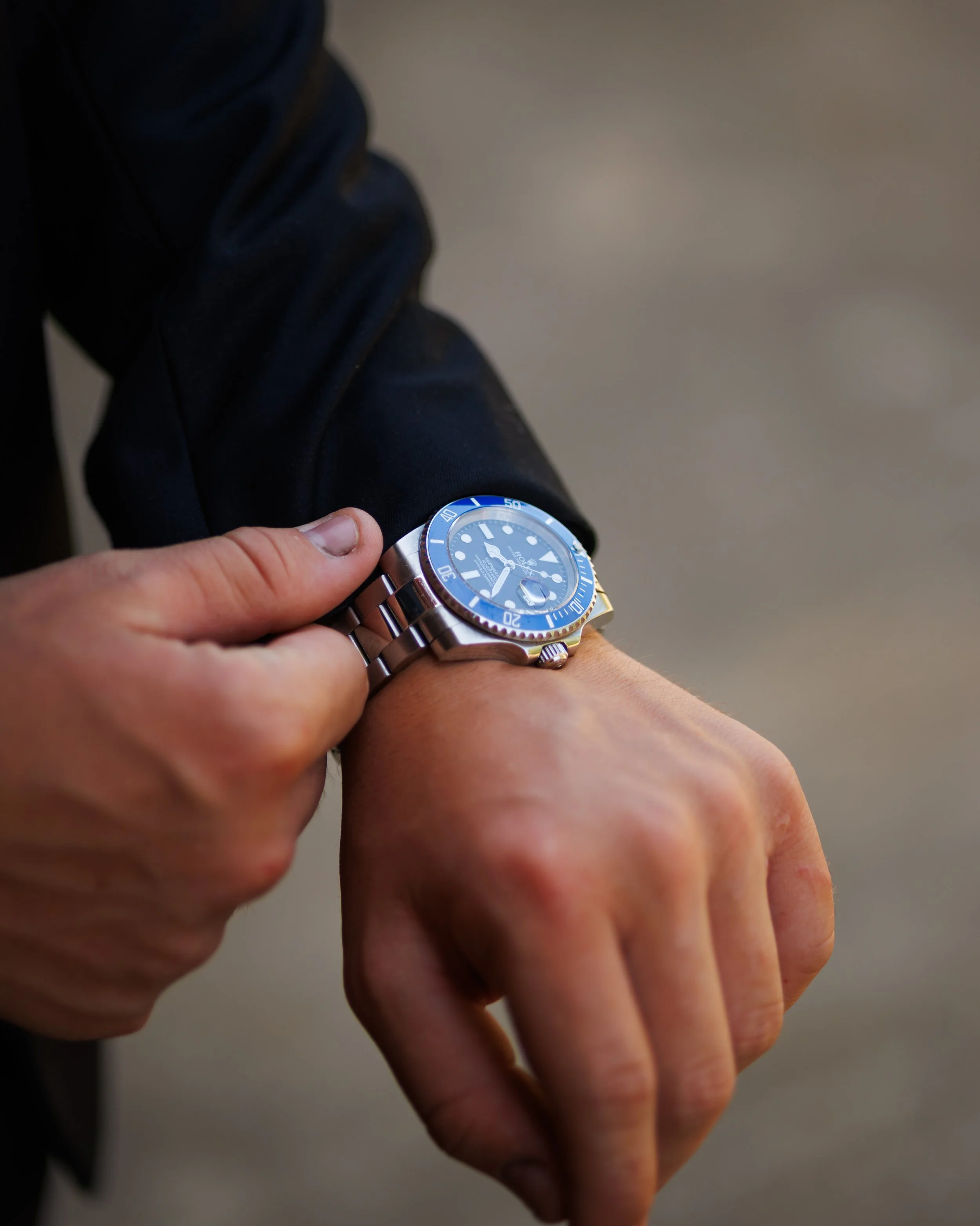 Close-up of a person's left wrist with a silver watch showing the time, on a person's left hand, with fingers and thumb visible, in front of another person's right hand, which is on their arm.
