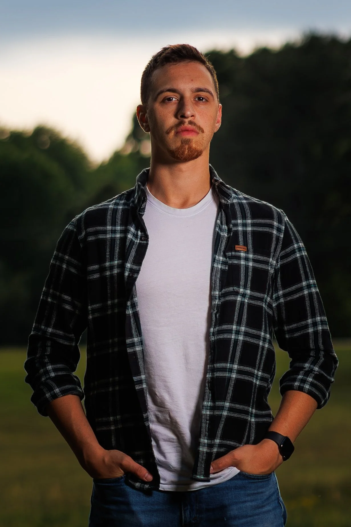 Young man standing outdoors in a casual pose, wearing a black and white checkered shirt over a white t-shirt, with his hands in his pockets, against a blurred natural background.