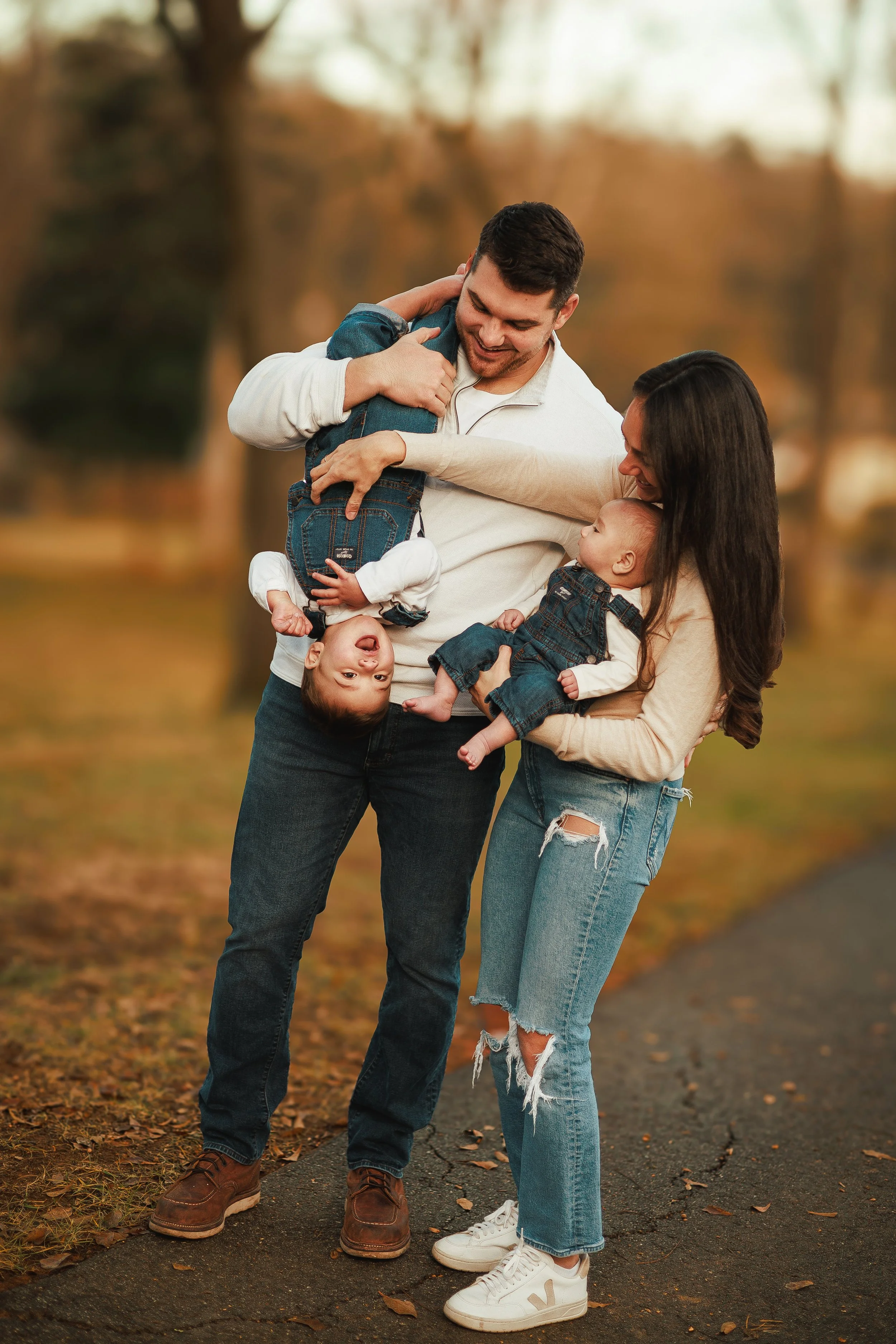 A happy family of four outdoors during fall, with trees and fallen leaves in the background. The father is holding a toddler upside down, both smiling. The mother is holding a baby, and they are all dressed casually.