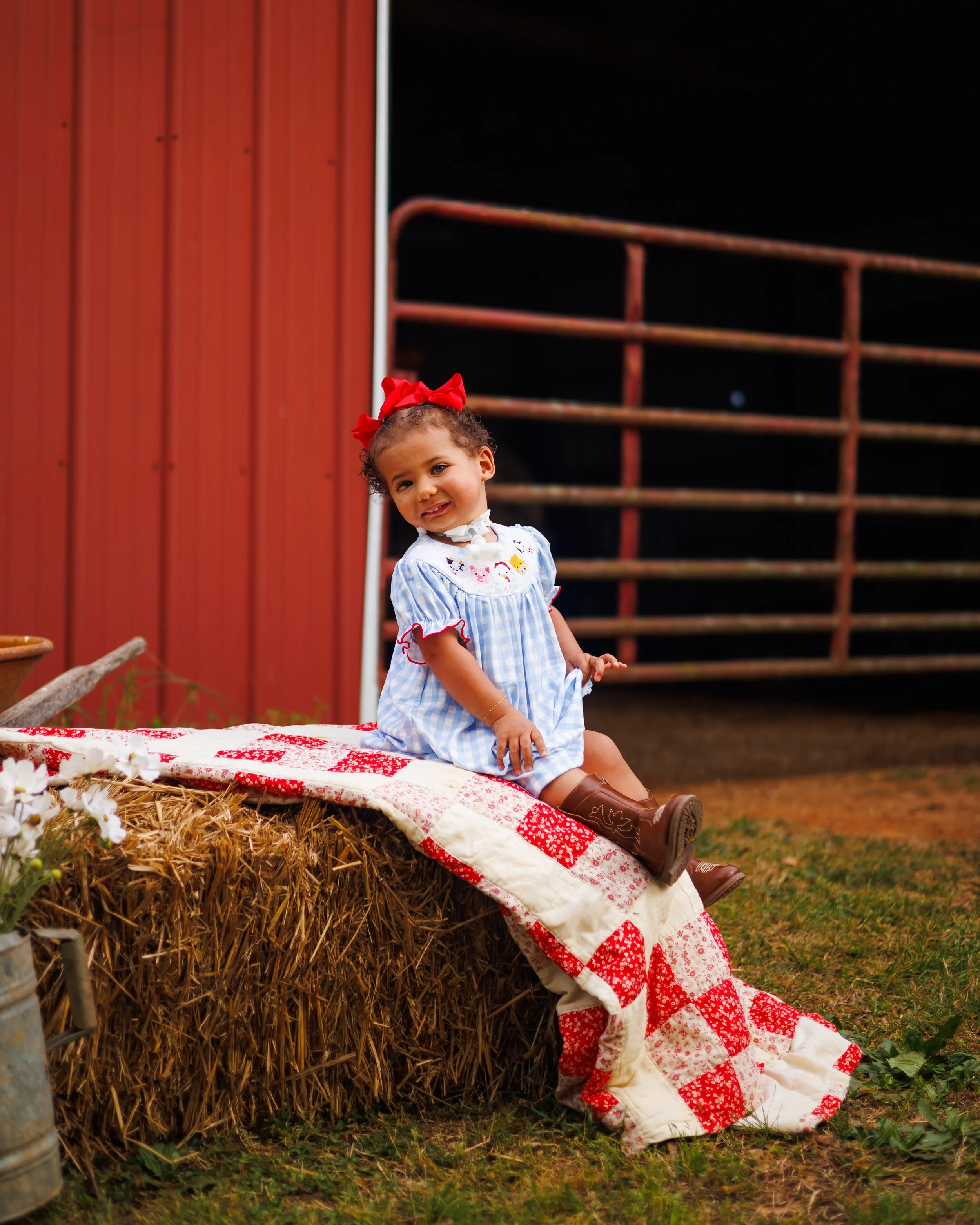 A young girl with curly hair, wearing a white and blue dress with red accents and a red bow, sitting on a quilt draped over a hay bale at night. There is a red barn and wooden structure in the background.