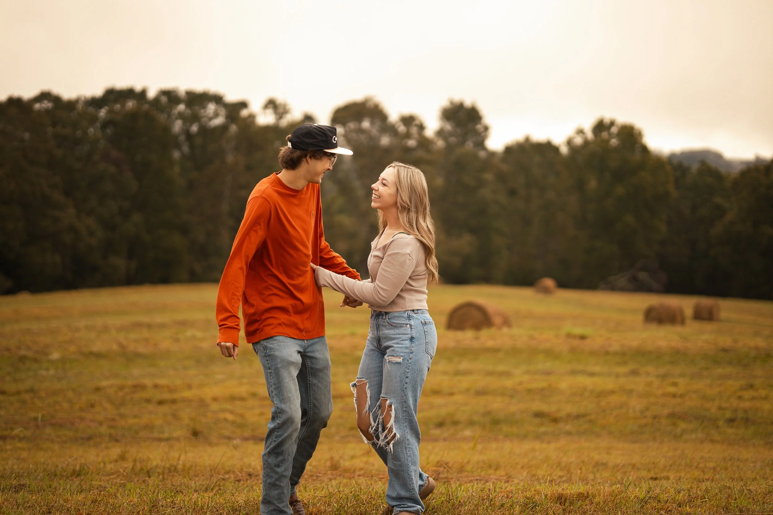 A young man and woman are holding hands and looking at each other, smiling, in an outdoor field with trees in the background during the daytime.