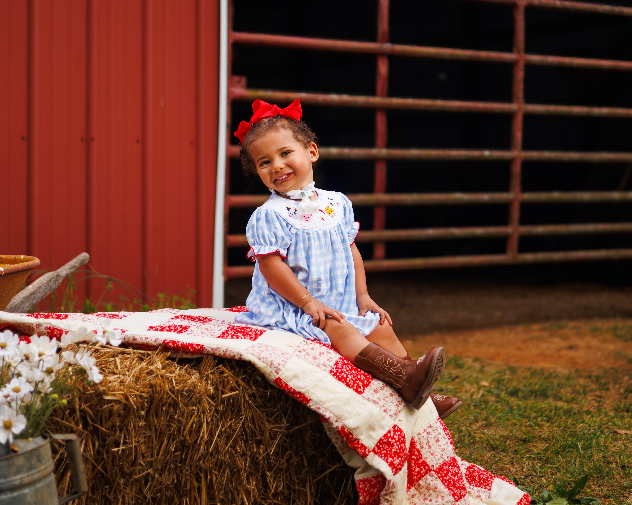 Young girl with curly hair, red bow, and cowboy boots, sitting on a hay bale in front of a red barn, smiling at the camera.