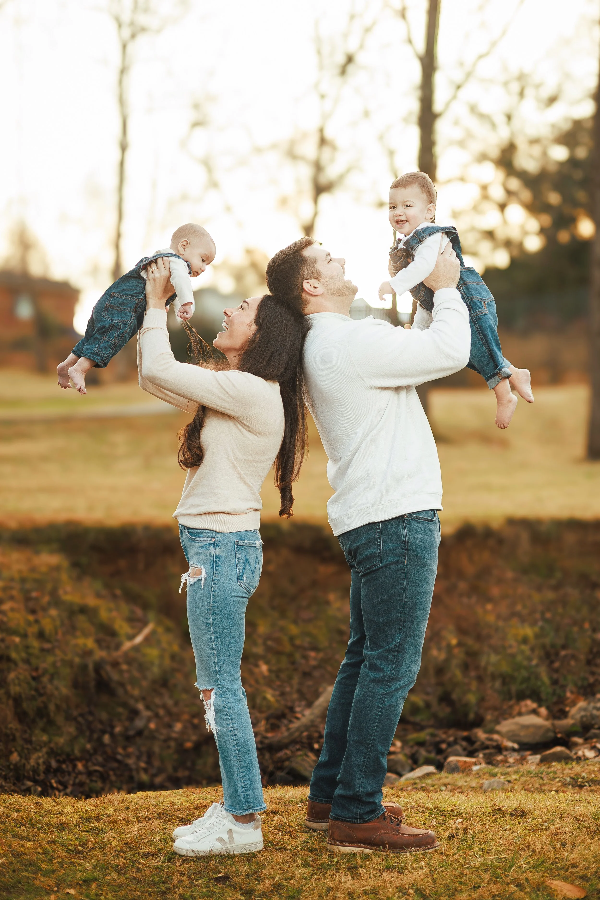 A family of four outdoors in a park during autumn. Two adults, a woman and a man, are holding their young children, a boy and a girl, up in the air. The woman with dark hair and ripped jeans supports the boy, while the man with short hair and a beard
