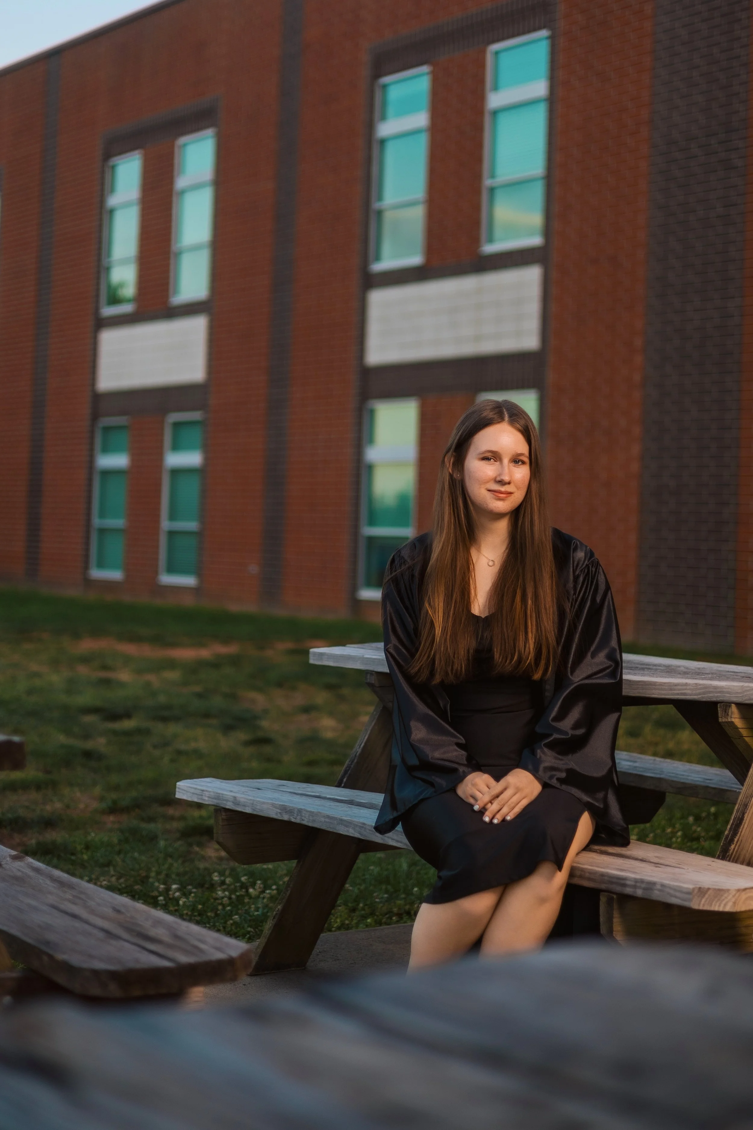 A young woman with long brown hair sitting on a wooden picnic table outdoors. She is wearing a black dress and a shiny black jacket. Behind her is a brick building with multiple windows. The scene appears to be at sunset or early evening.