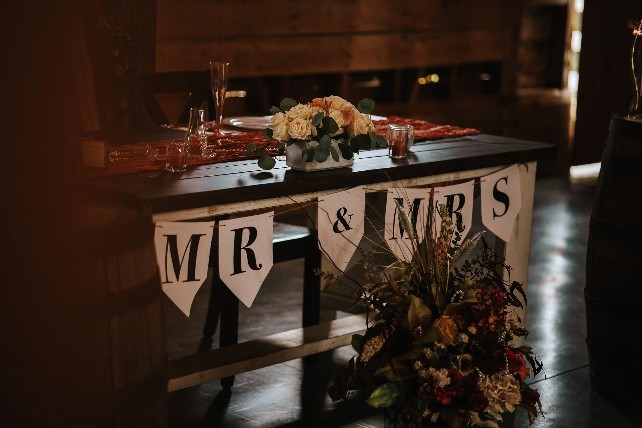 Wedding reception table decorated with a bouquet of white and peach roses in a white vase, candles, glassware, and a red table runner. There are 'MR & MRS' banners hanging below the table. The setting is rustic with wooden walls and a cozy ambiance.
