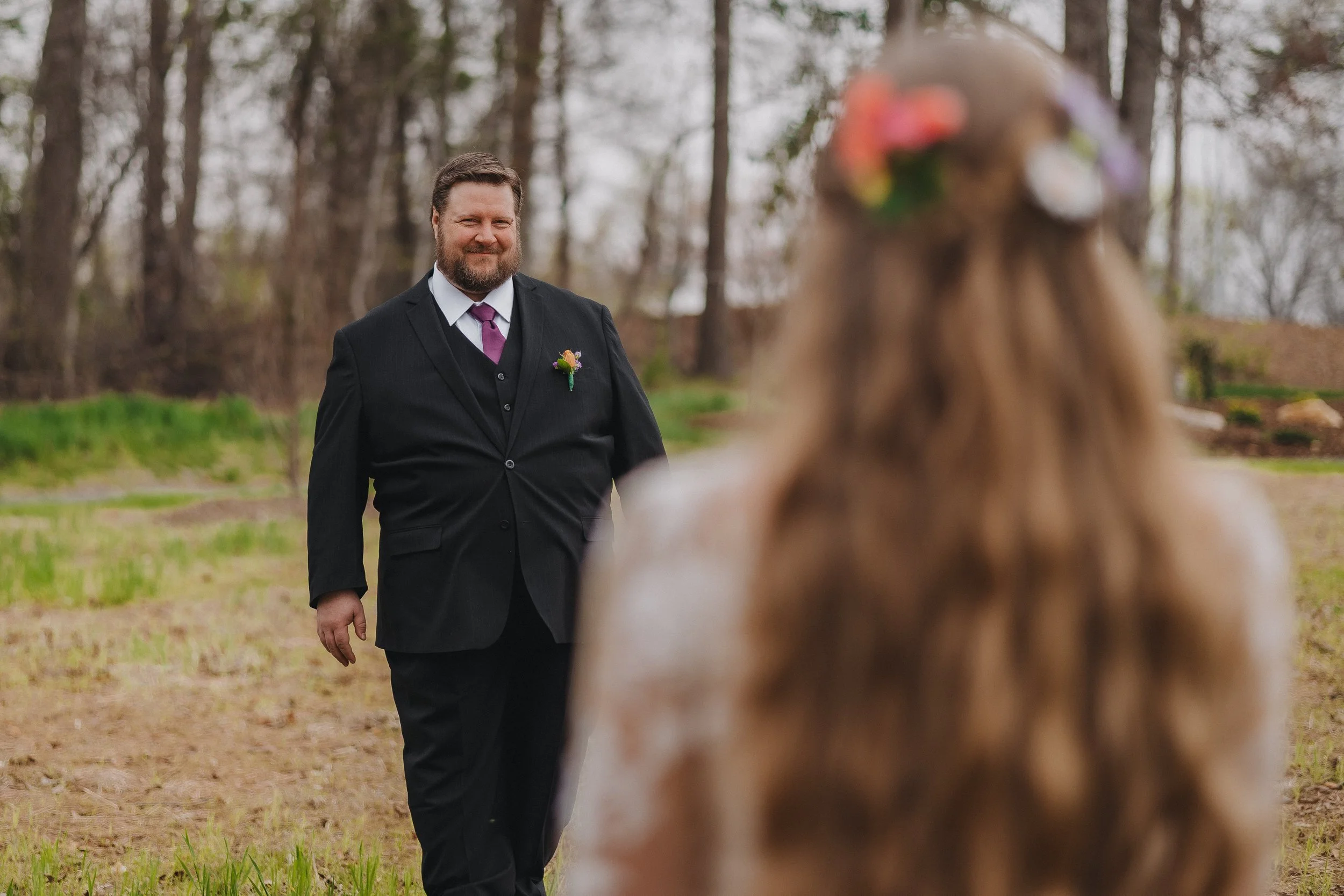 Man in a suit smiling at a dog in a park with trees.