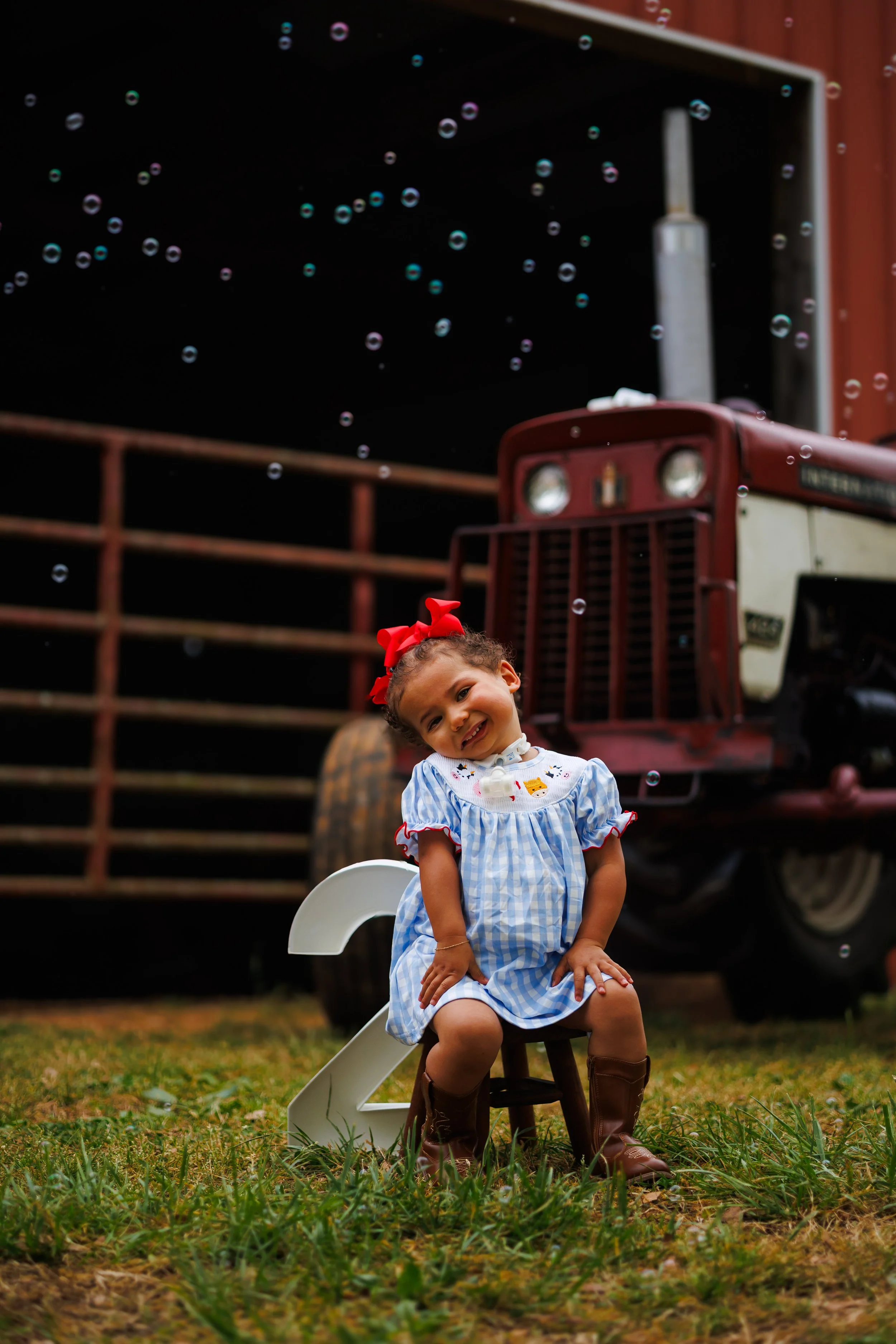 A young girl in a blue and white plaid dress with a red bow in her hair sitting on a small wooden chair outdoors, celebrating the New Year with large white numbers '2023' and a decorative tractor in the background, with floating bubbles in the air.