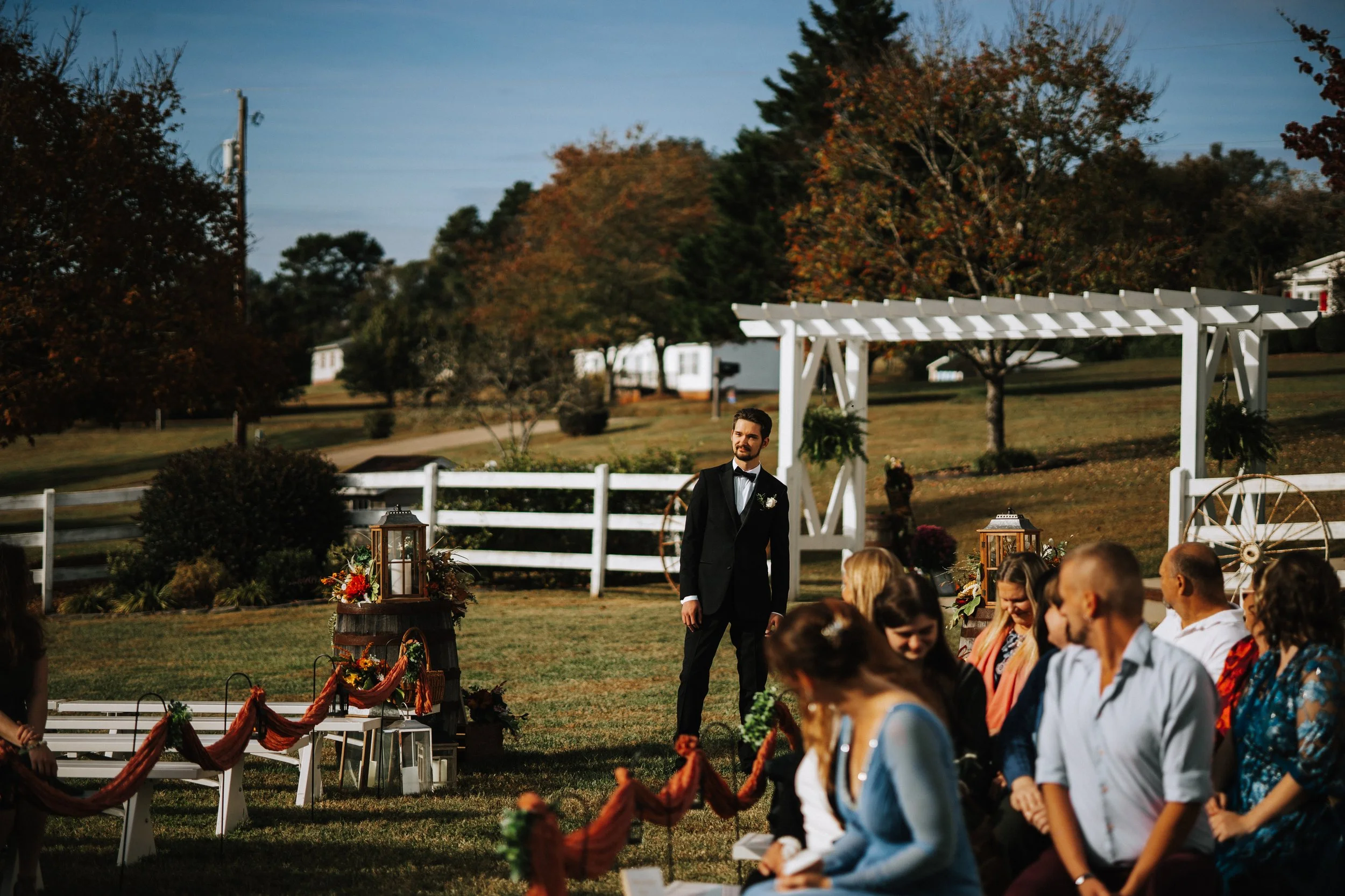 A groom in a black tuxedo stands during an outdoor wedding ceremony with seated guests around him. The setting features trees with fall foliage, white fencing, and decorative lanterns, under a clear blue sky.