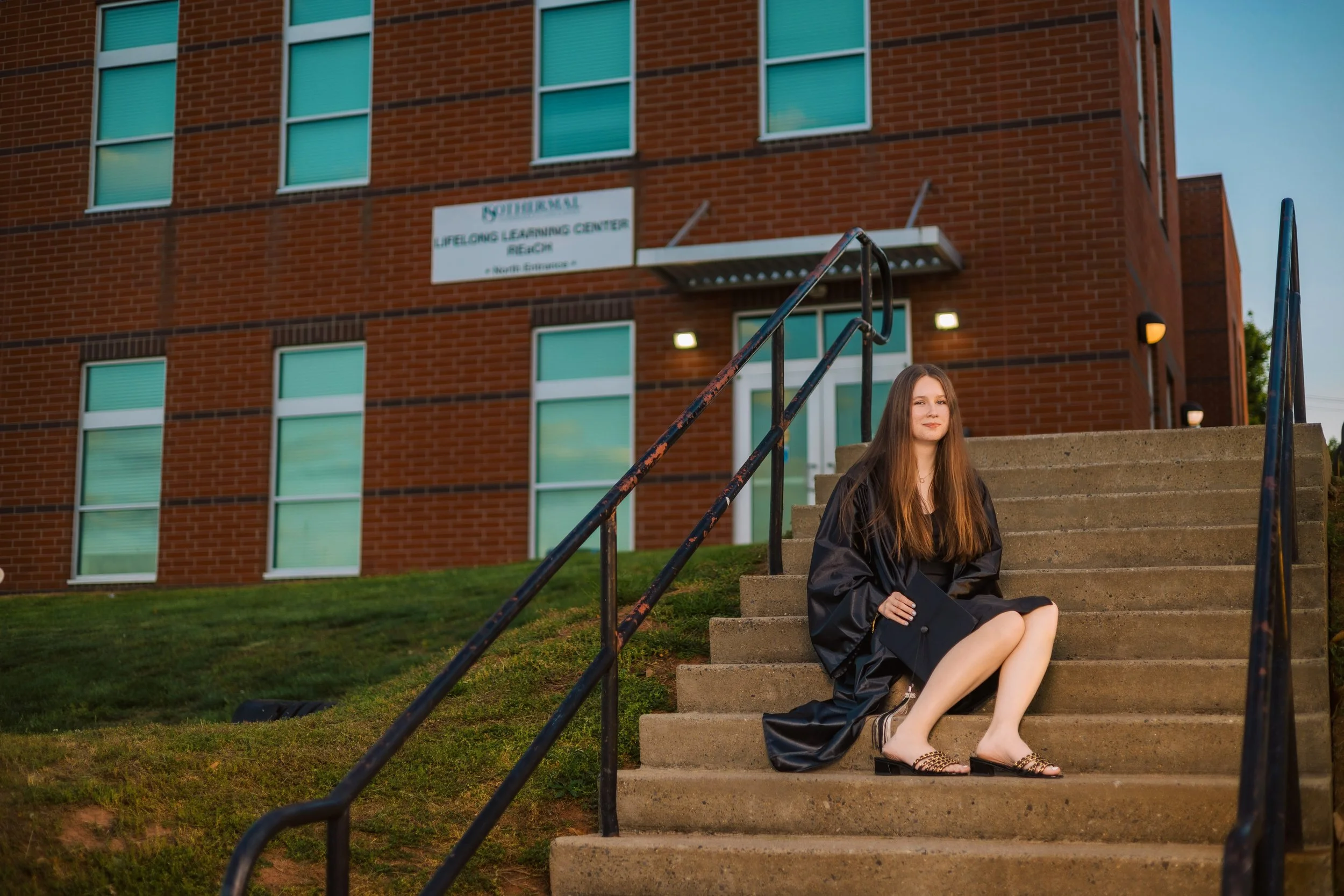 A young woman with long hair sitting on the steps outside a brick building, wearing a black graduation gown and holding a diploma, during sunset.