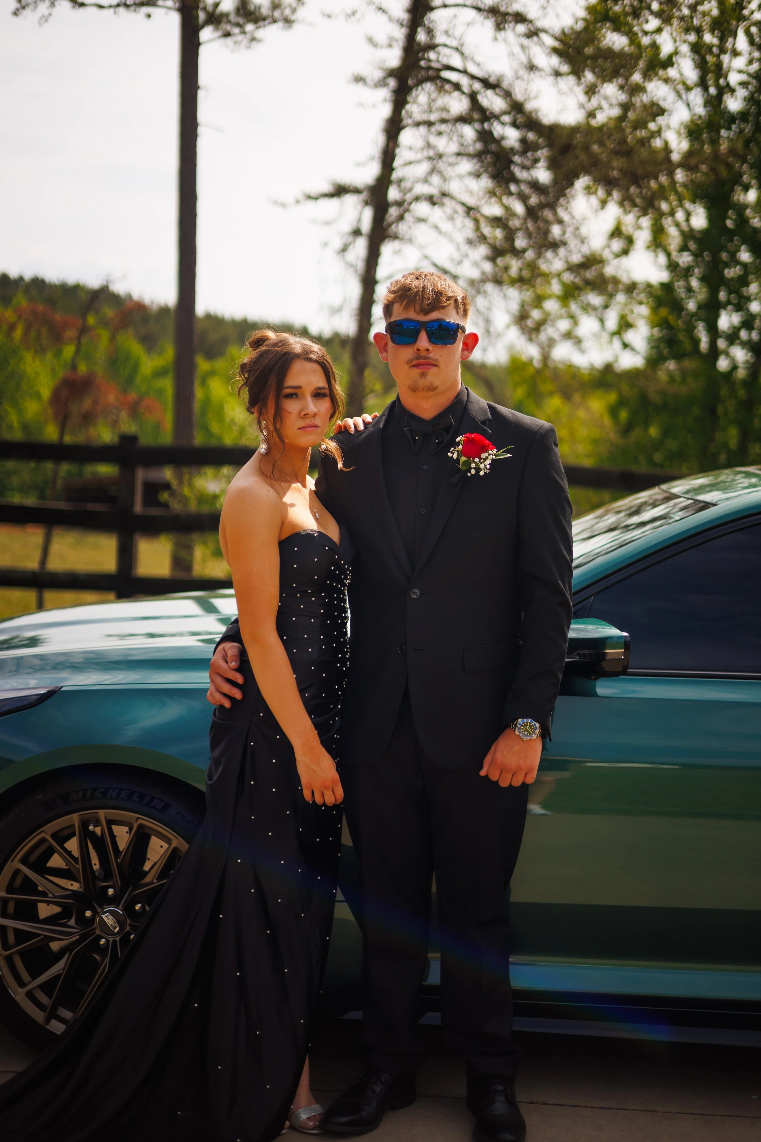 A young woman in a black evening gown and a young man in a black tuxedo with sunglasses pose together outdoors in front of a teal luxury car, with trees and a wooden fence in the background.