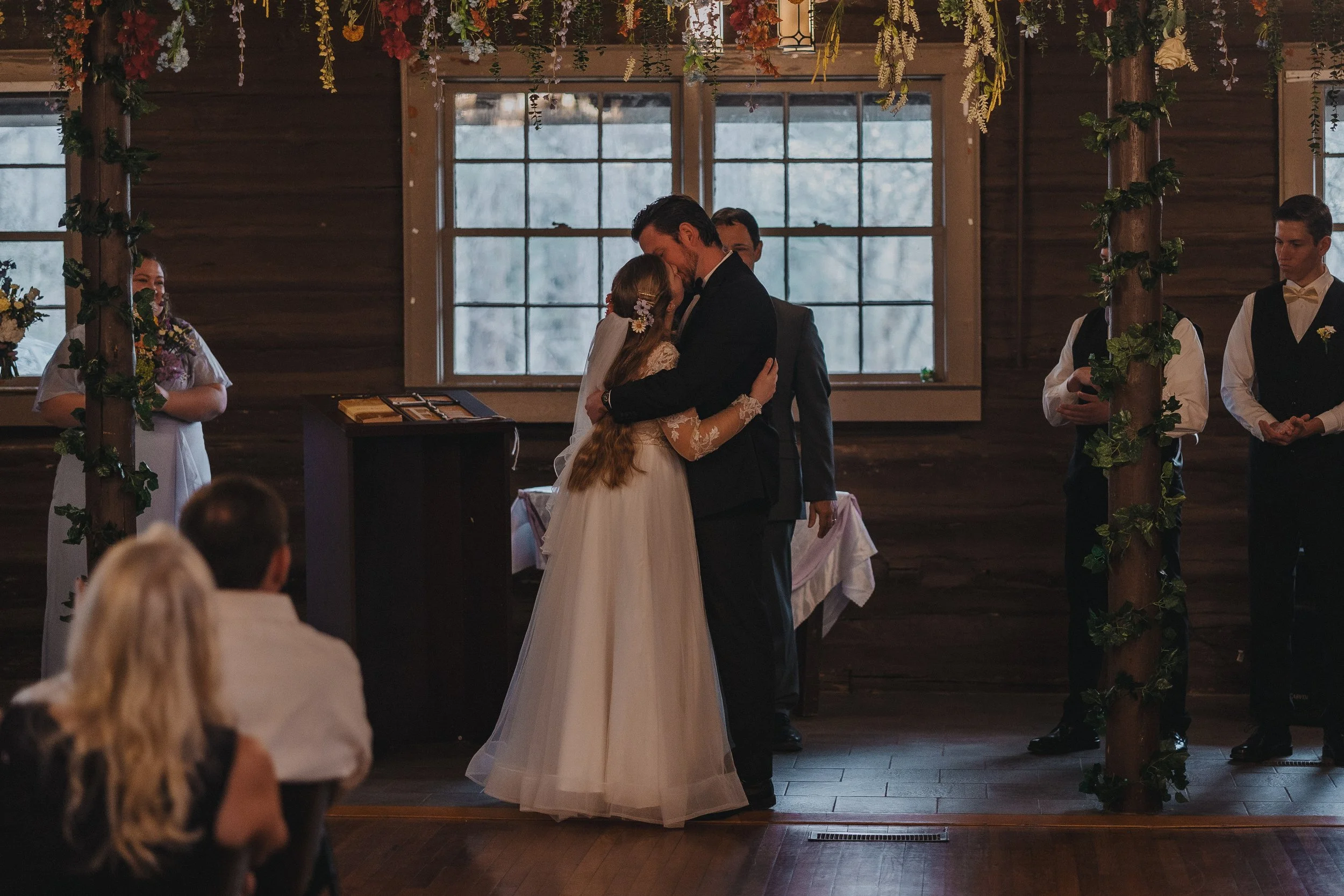 A bride and groom share a kiss during their wedding ceremony inside a rustic wooden chapel decorated with hanging flowers and greenery, with guests seated watching.