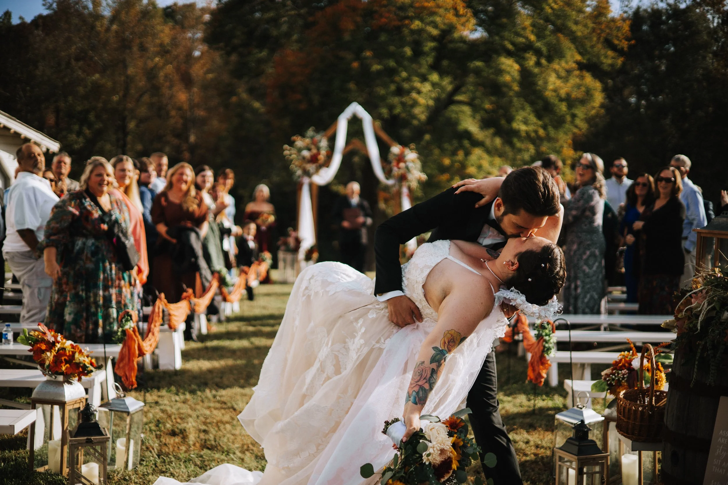 A couple dances at an outdoor wedding ceremony, with guests smiling and watching in the background, decorated with fall-themed flowers and lanterns.
