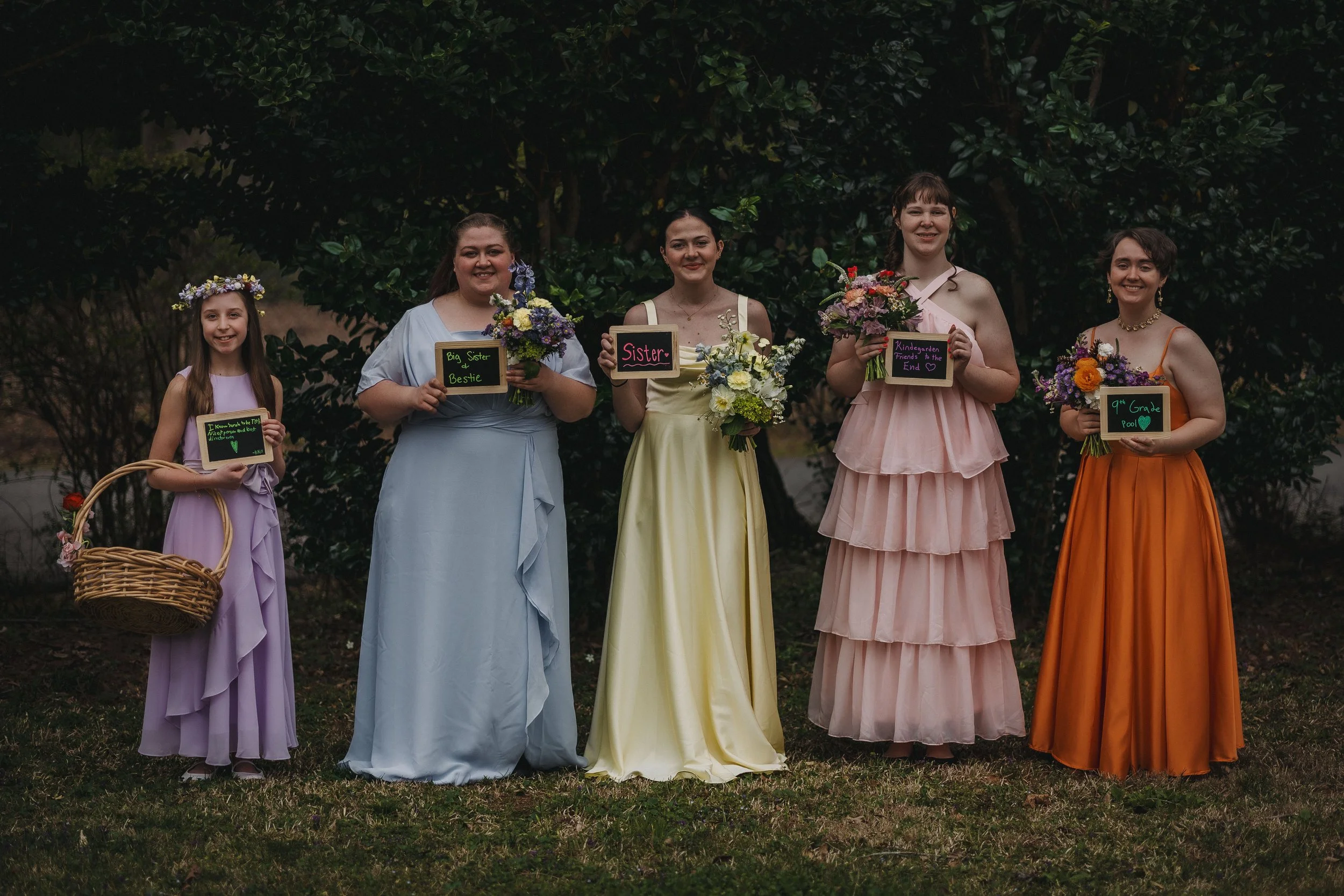 Group of five women of different ages standing in a line outdoors, dressed in pastel and bright colored dresses, holding flowers and small chalkboard signs with messages, with greenery and trees in the background.