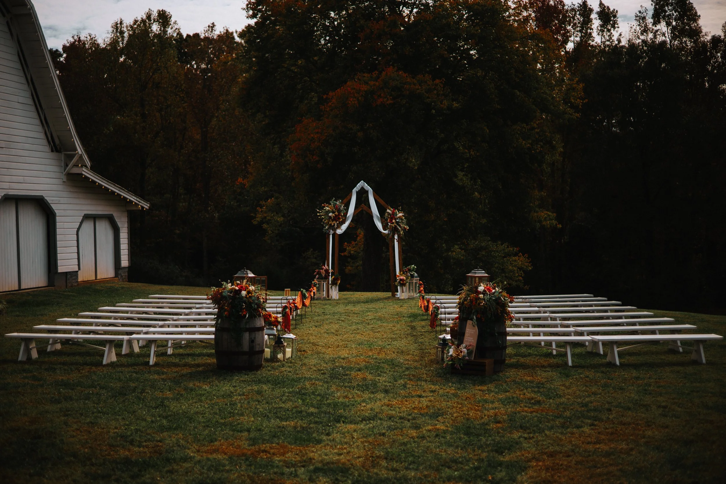 Outdoor wedding ceremony setup with white benches, floral arrangements, and a decorated arch, surrounded by trees with autumn leaves.