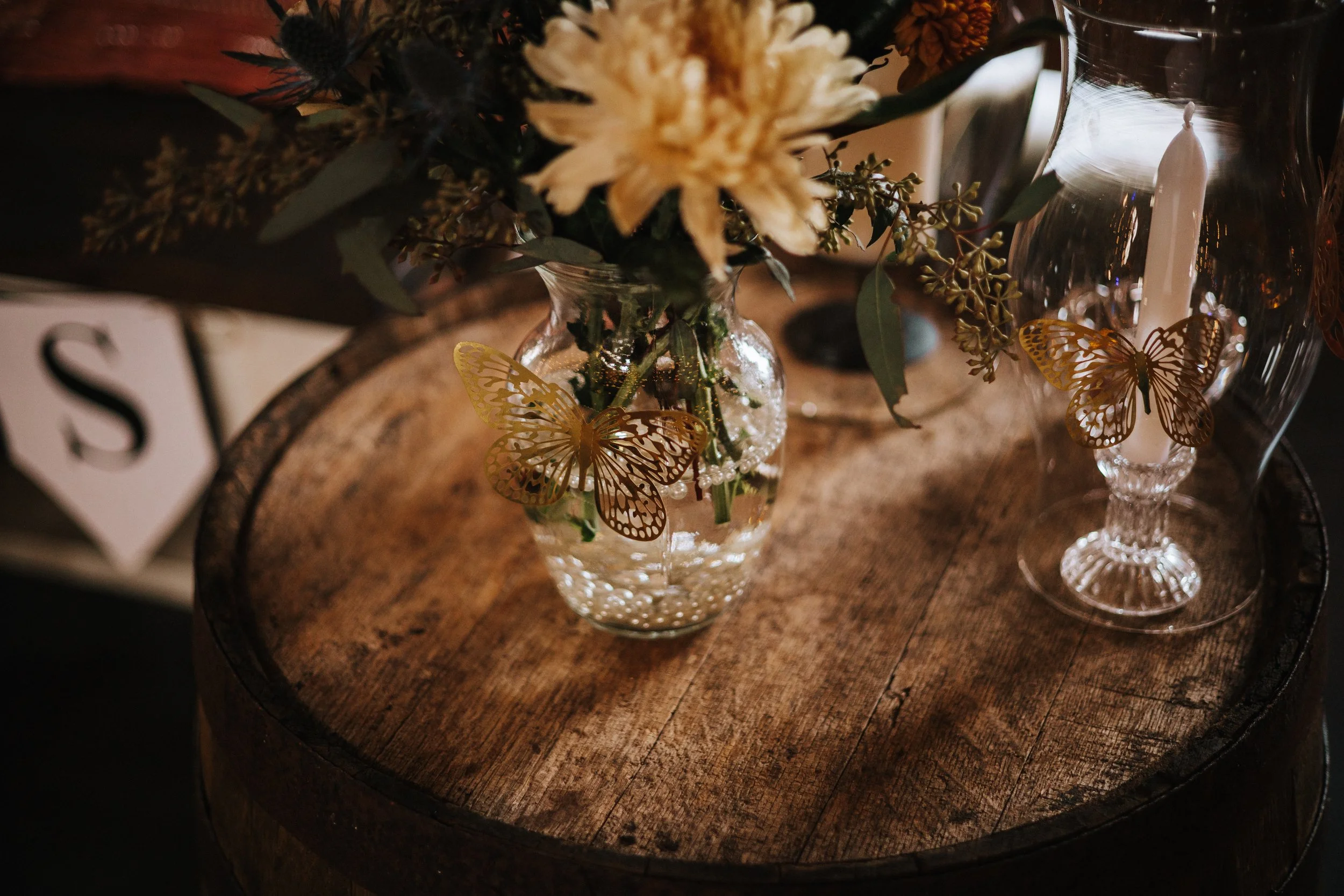 A glass vase with a floral arrangement including white flowers and greenery, decorated with decorative gold butterfly ornaments, placed on a round wooden table with a candle in a glass holder nearby.