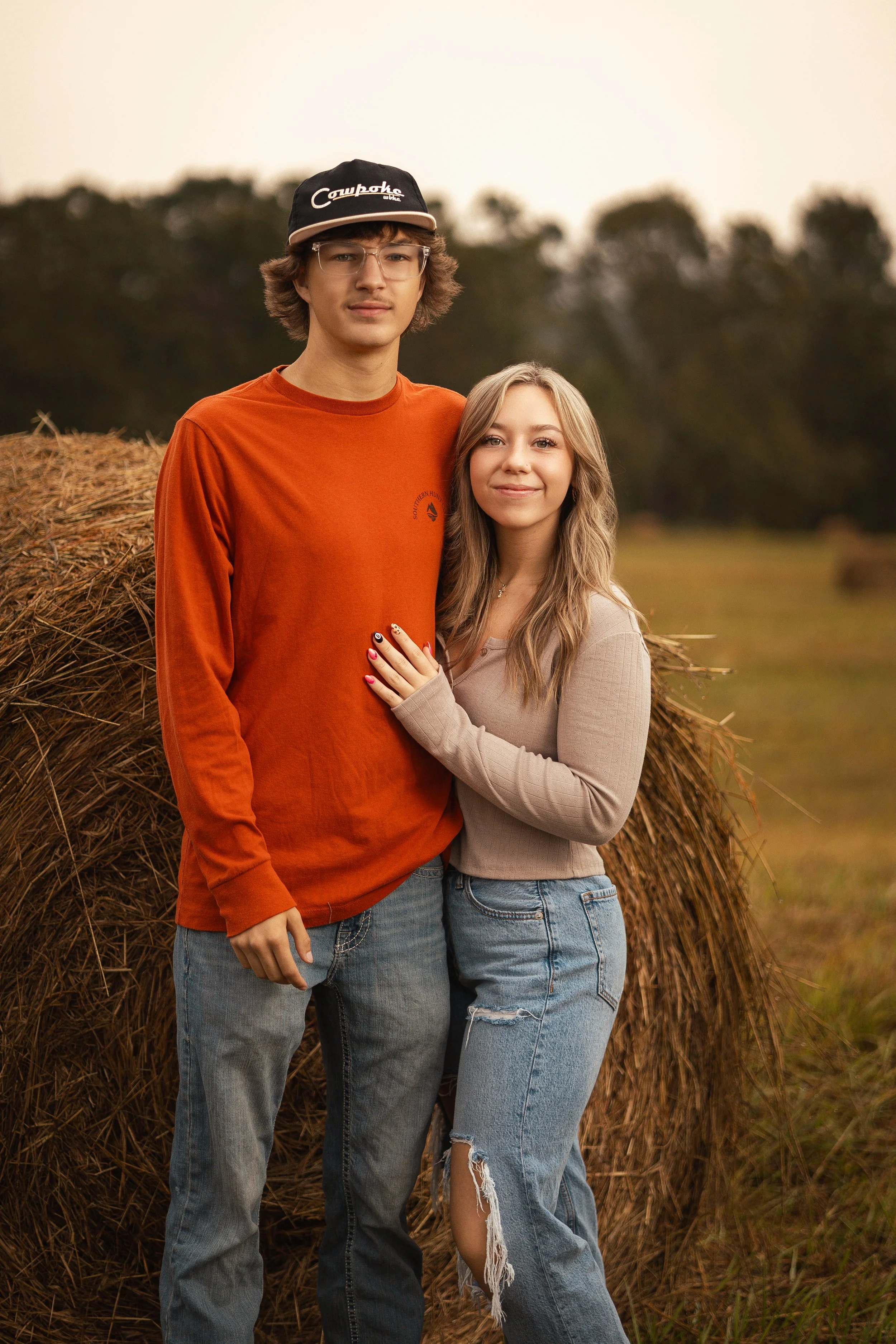 A young man and woman standing in front of a large hay bale outdoors, with trees in the background during sunset.
