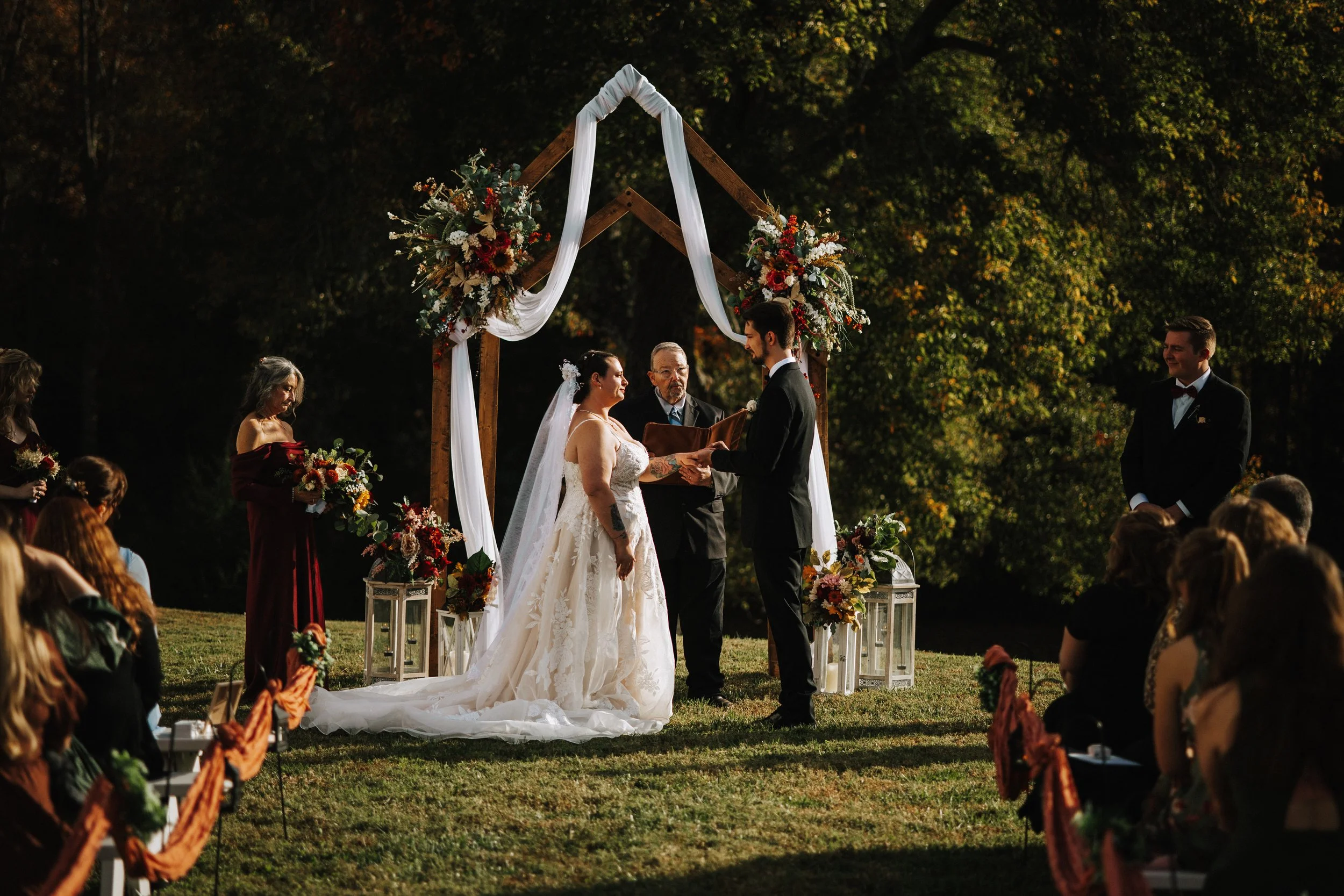 A wedding ceremony taking place outdoors under a wooden arch decorated with flowers and white fabric, with a bride and groom exchanging vows, surrounded by guests seated on either side and greenery in the background.