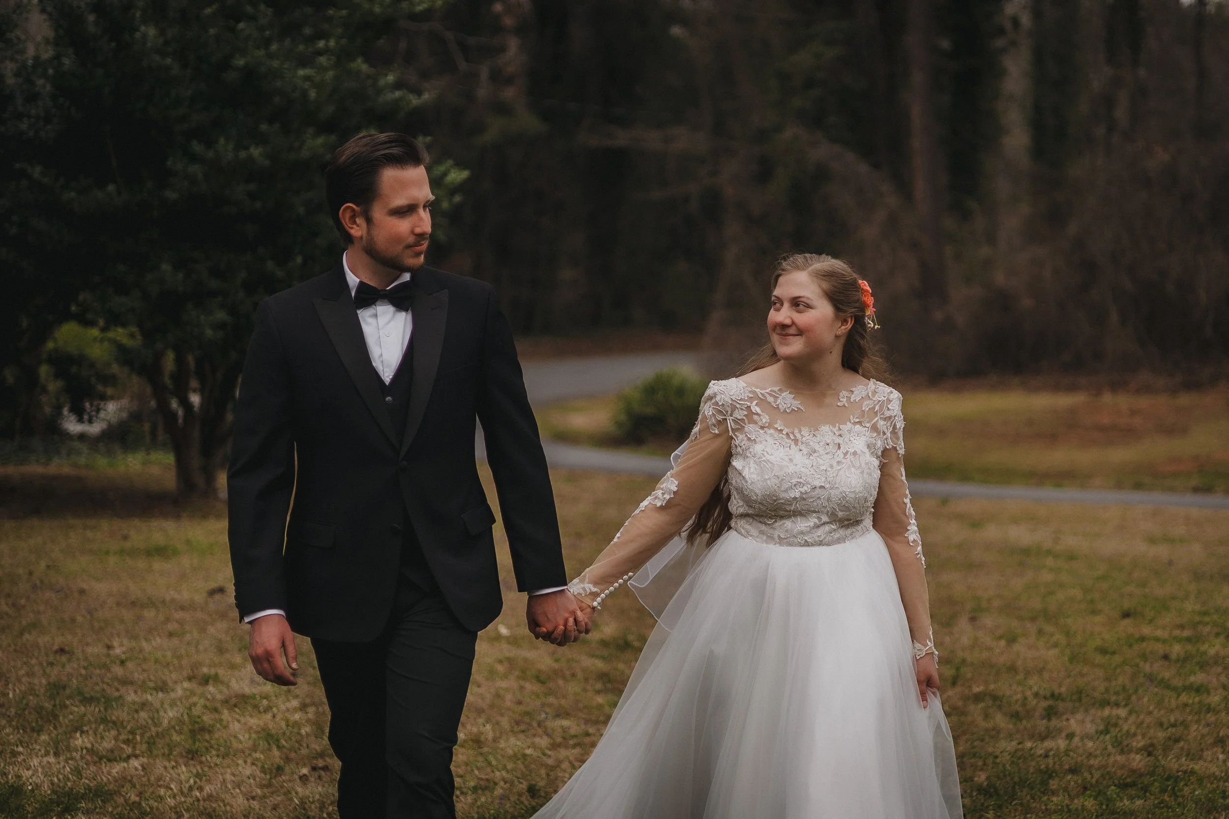 A bride and groom holding hands outdoors, walking on grass with trees in the background; the bride is wearing a white lace wedding gown with long sleeves and floral hair accessories, the groom is in a black tuxedo with a bow tie.
