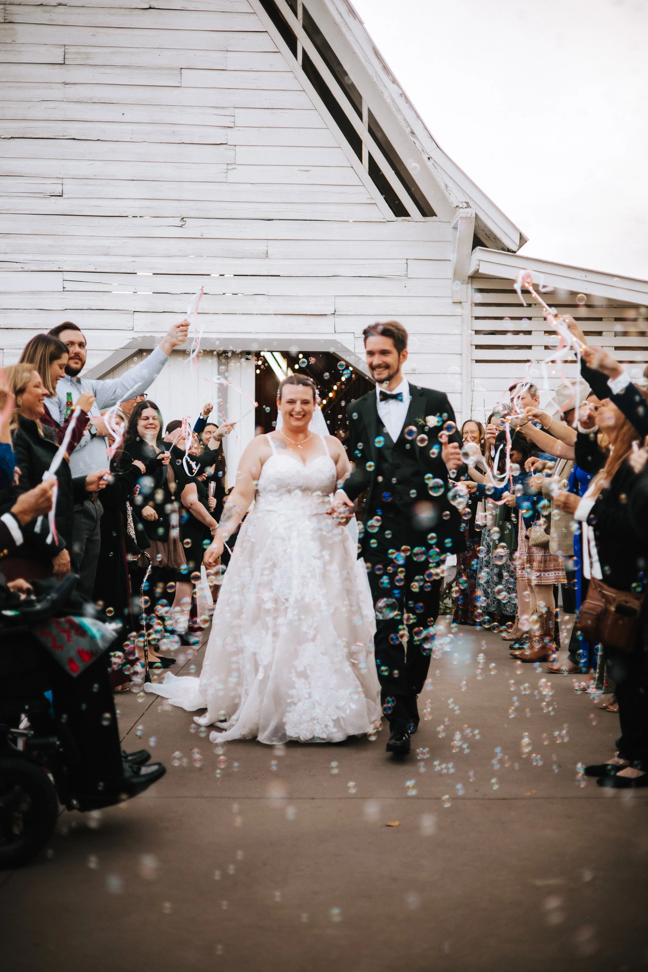 A bride and groom walking through a crowd of people celebrating with bubbles and confetti outside a white wooden building.