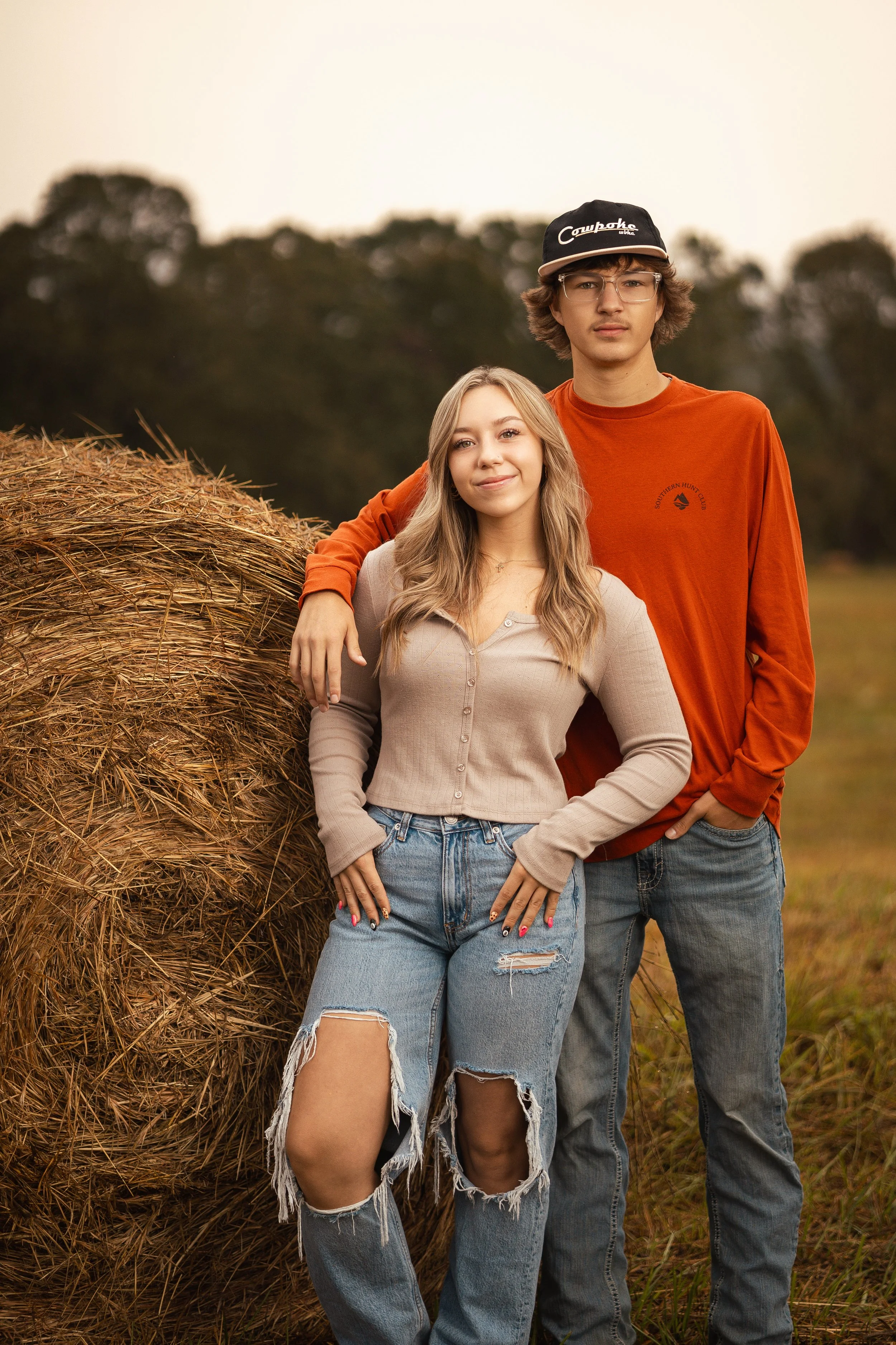 A young woman and young man standing outdoors at sunset, next to a large hay bale. The woman has long blonde hair, a beige button-up shirt, and ripped jeans. The man has brown hair, glasses, an orange long-sleeve shirt, and a black cap. Both are look
