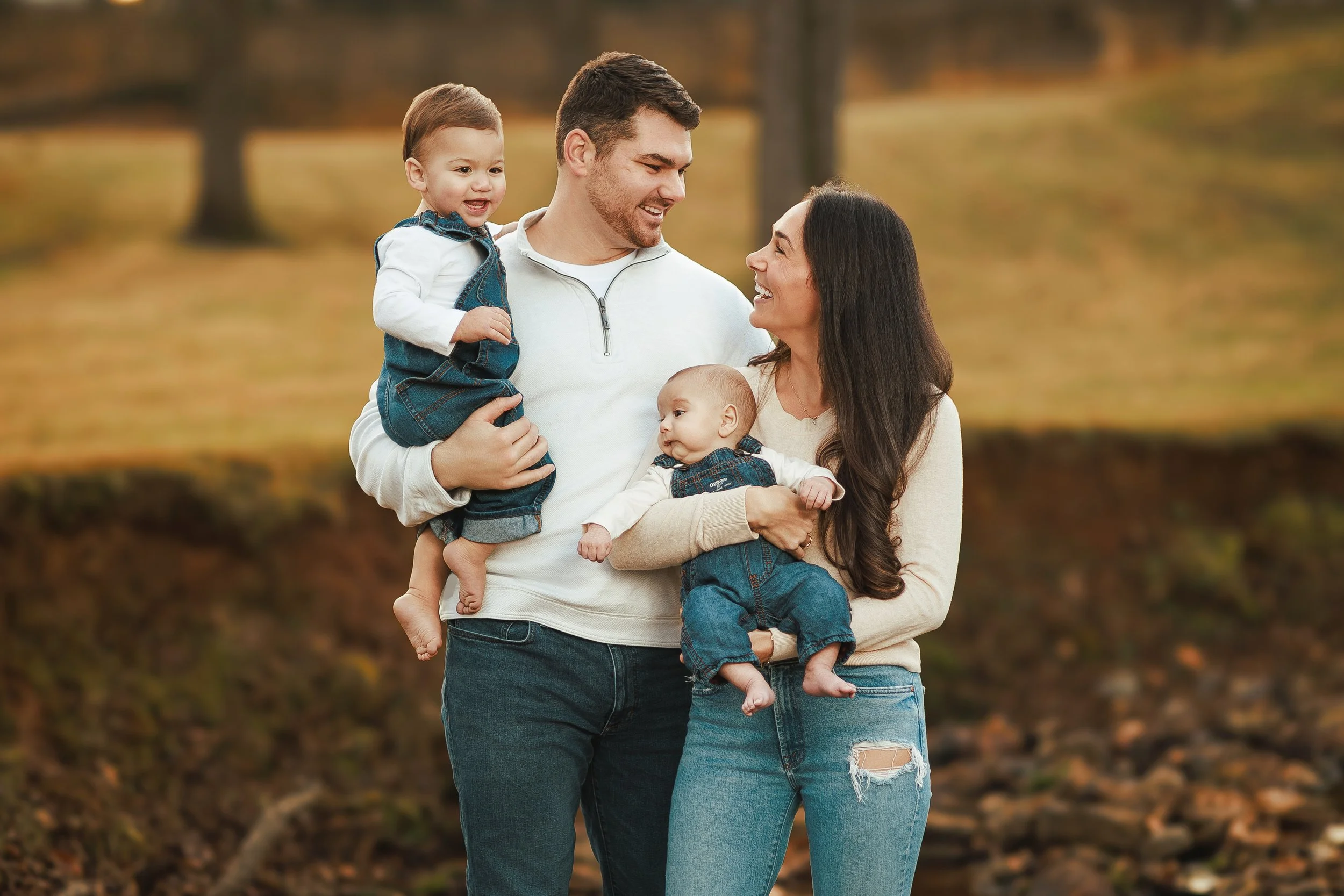 A happy family of four, including a man, woman, and two young children, enjoying an outdoor fall setting with trees and fallen leaves in the background.