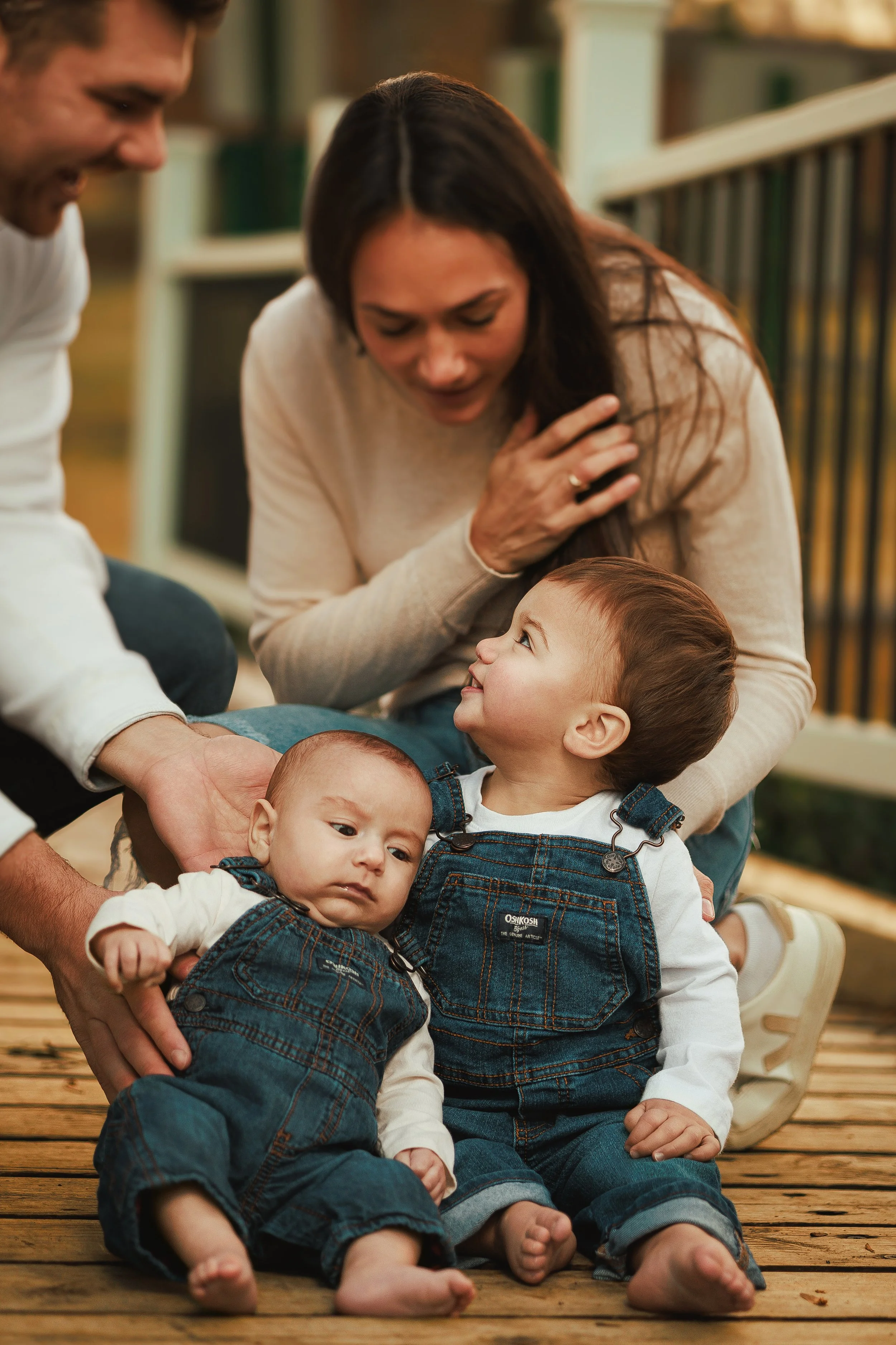 Two women and three children sitting on a wooden deck with bookshelves in the background. The women are smiling and the children are lying on their laps, engaging with each other.