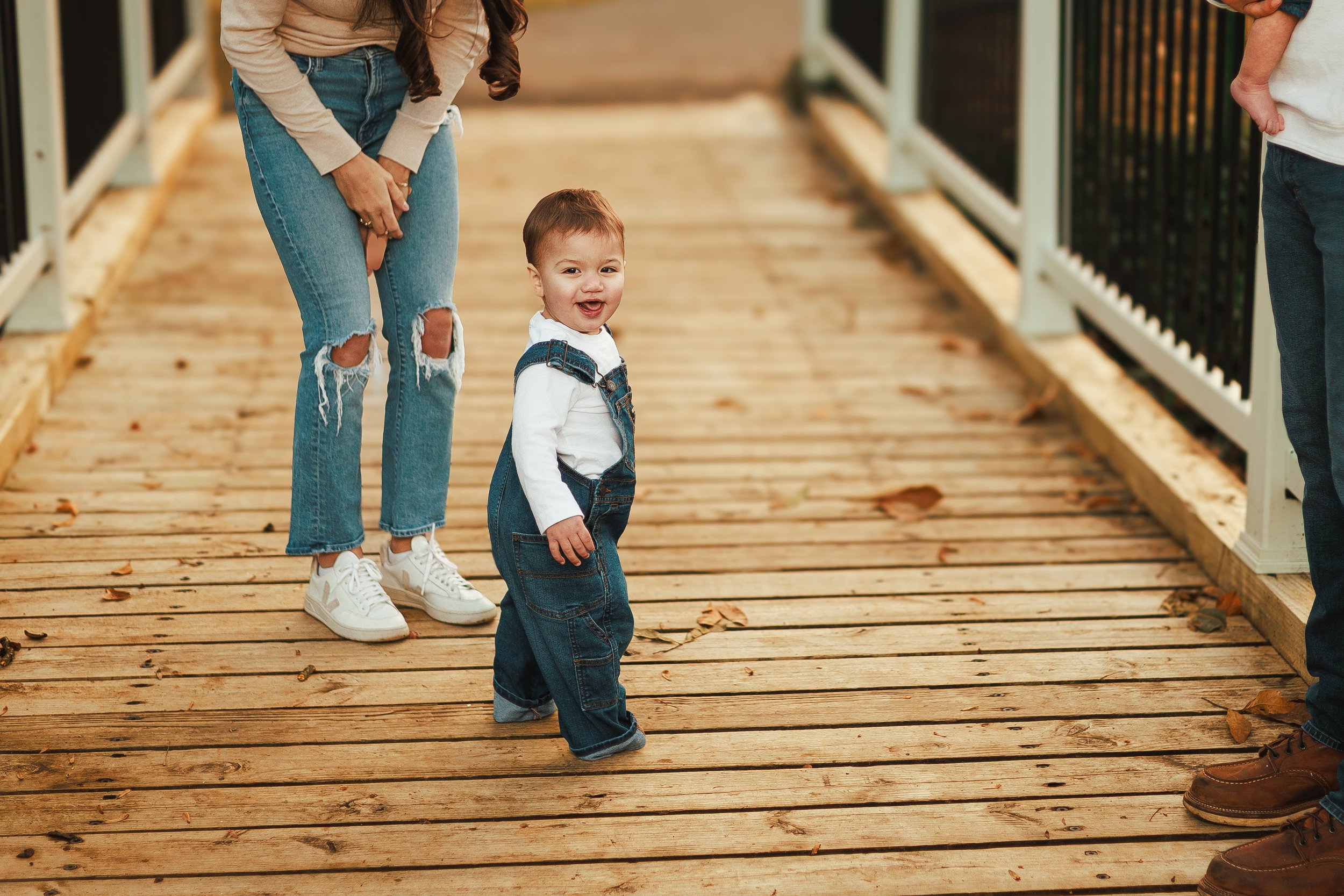 A smiling young boy in denim overalls and a white shirt standing on a wooden bridge, with two adults, one on each side, partially visible. The woman on the left is wearing ripped jeans and white sneakers, while the man on the right is wearing dark je