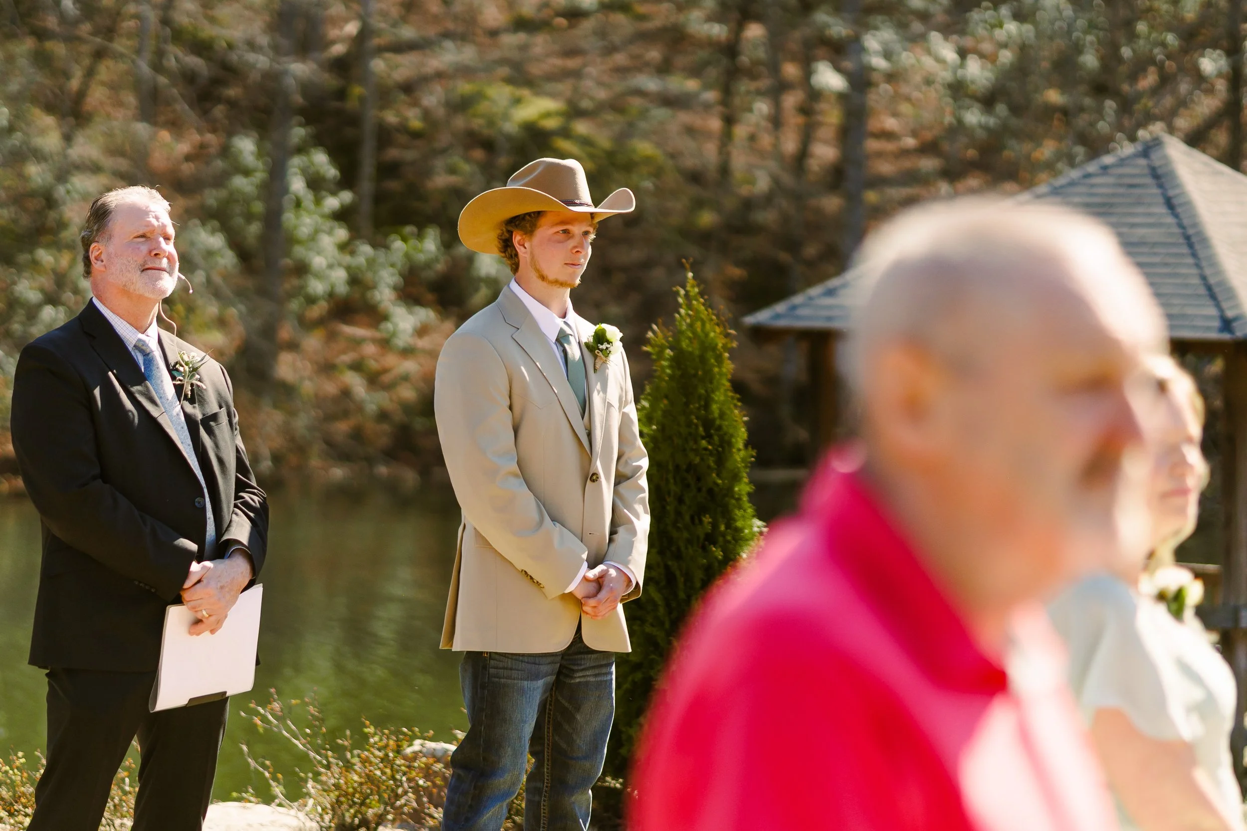 Three men standing outdoors during a wedding ceremony, with autumn trees in the background. One man is in a business suit, another in a cream-colored suit with a cowboy hat, and the third is partially visible in the foreground wearing a pink jacket.