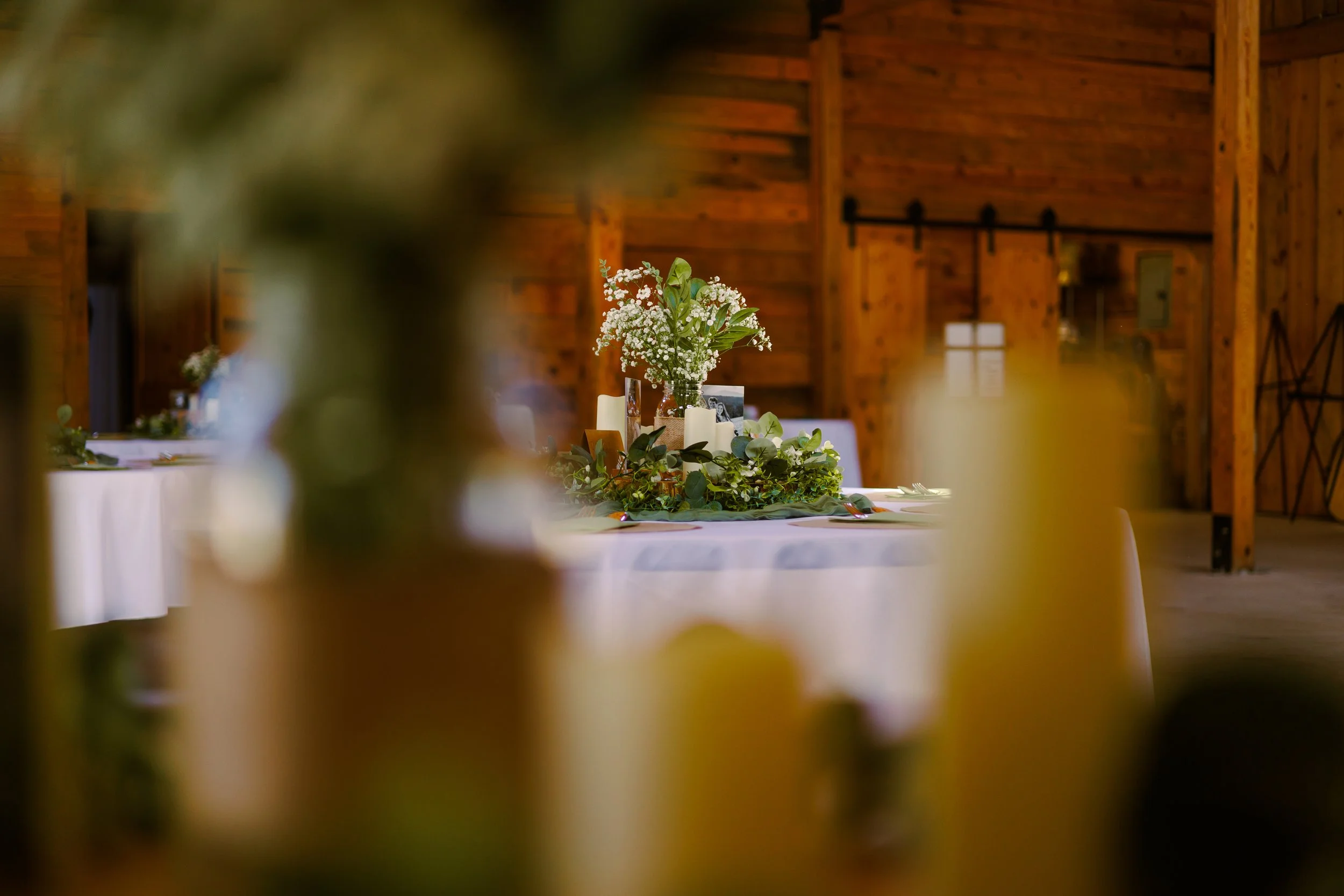 A decorated table with a floral centerpiece, candles, and framed photos inside a rustic wooden barn.