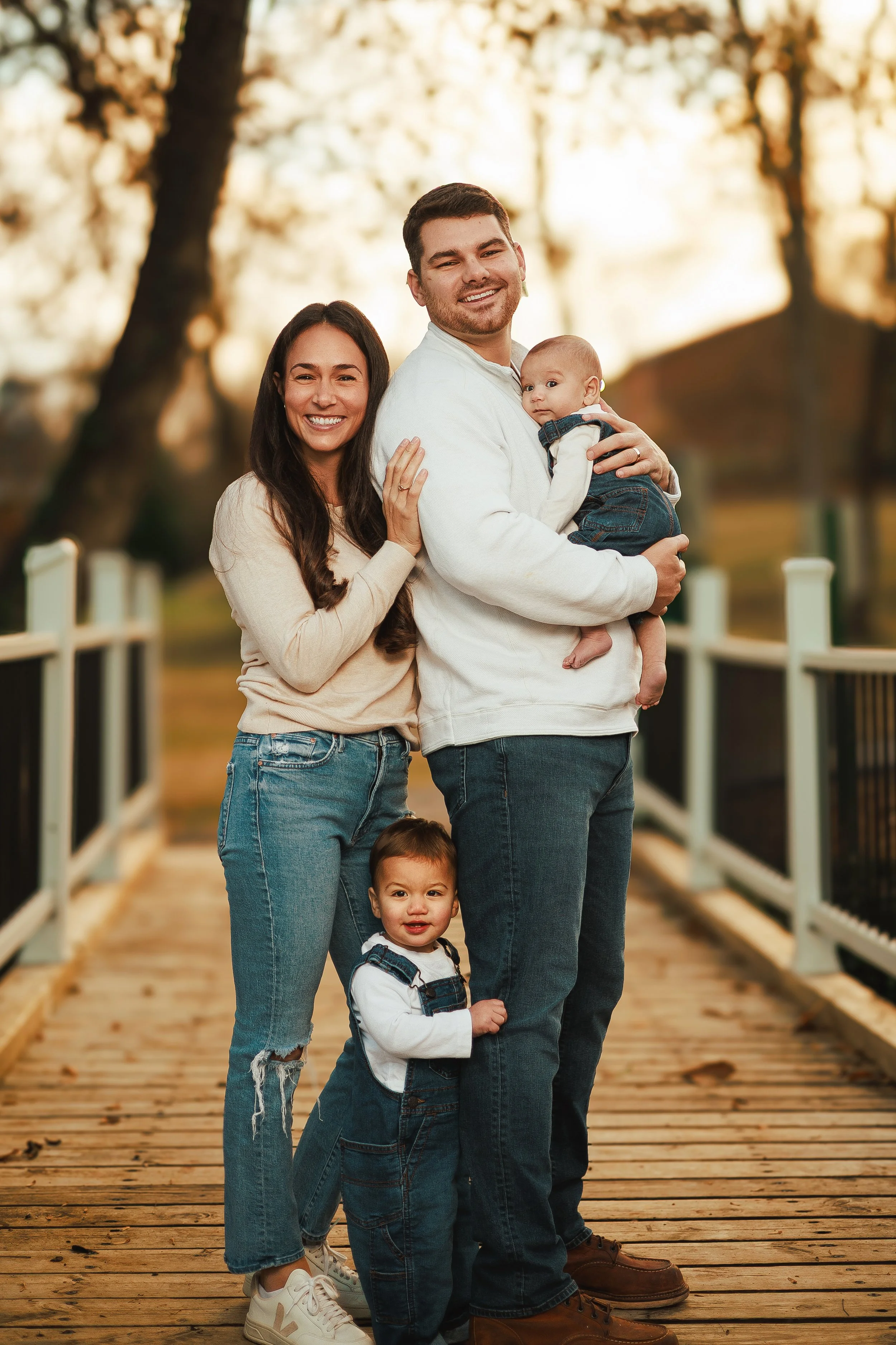 A family of five standing on a wooden bridge outdoors during autumn, with trees and golden leaves in the background. The family includes a woman with long dark hair, a man, two young boys, and a baby.