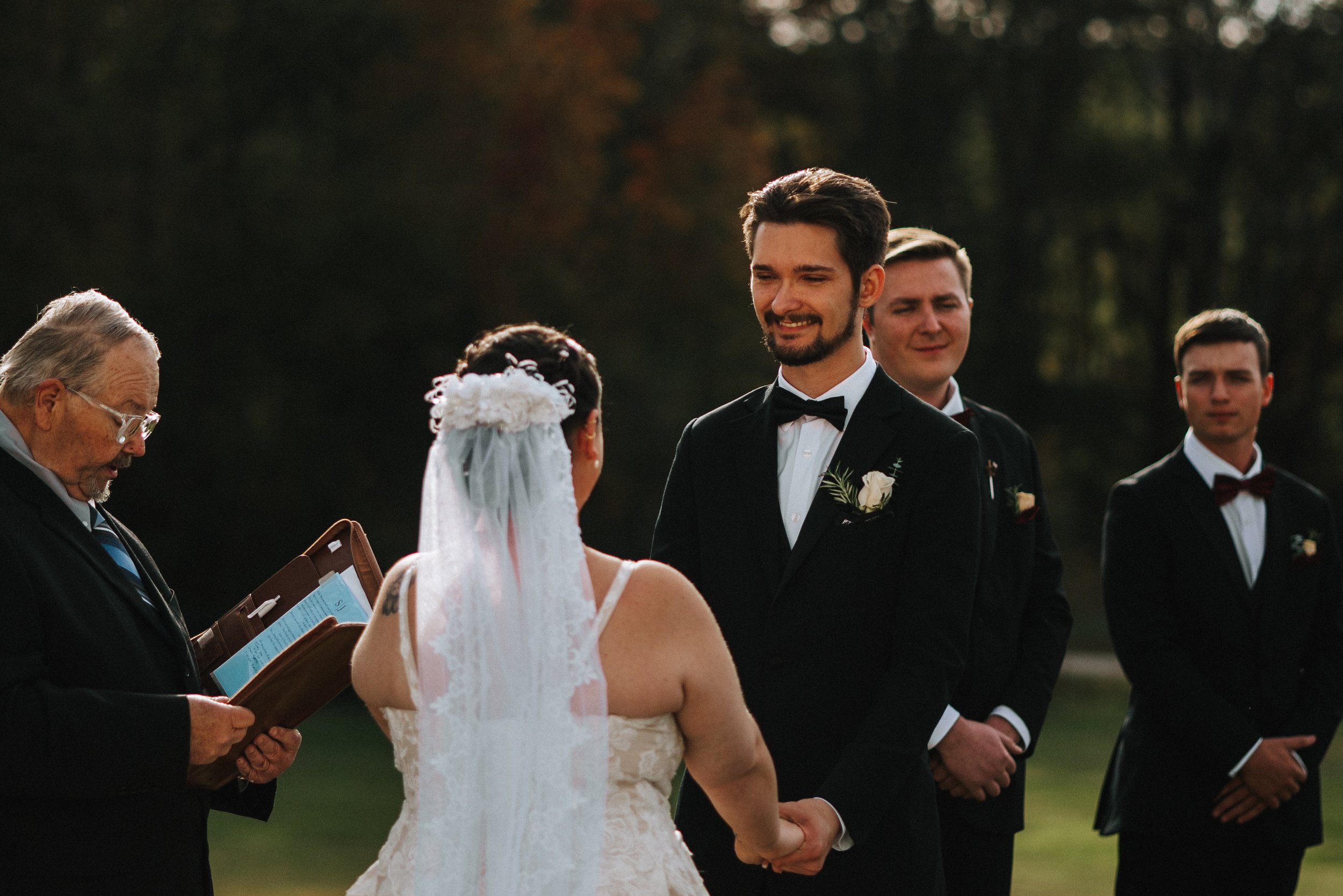 Couple exchanging vows during outdoor wedding ceremony, with officiant reading and officiant and groomsmen standing in formal attire.