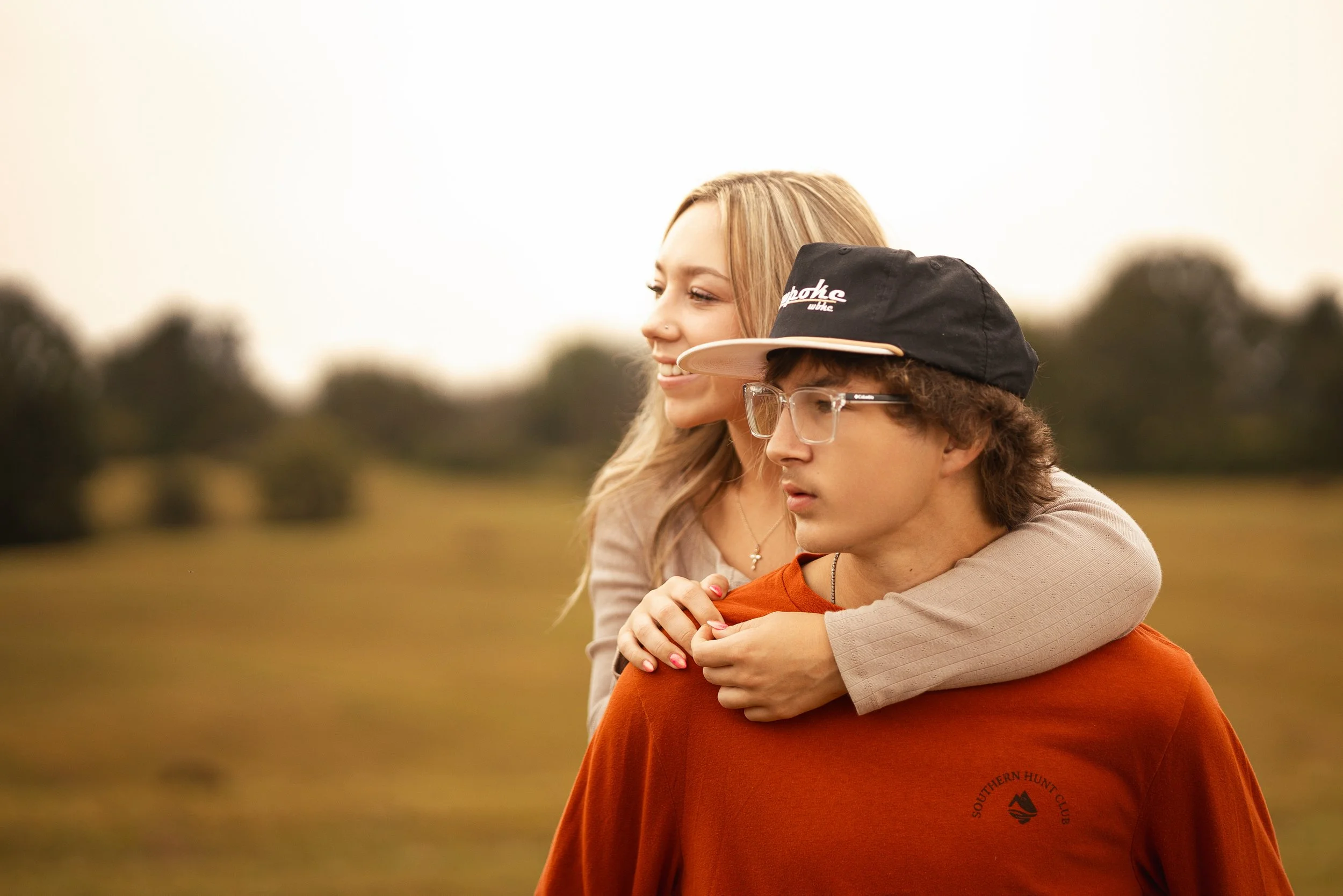 Two people sharing a hug outdoors, one with long blonde hair and the other with short brown hair, wearing glasses and a cap, in a park or field.