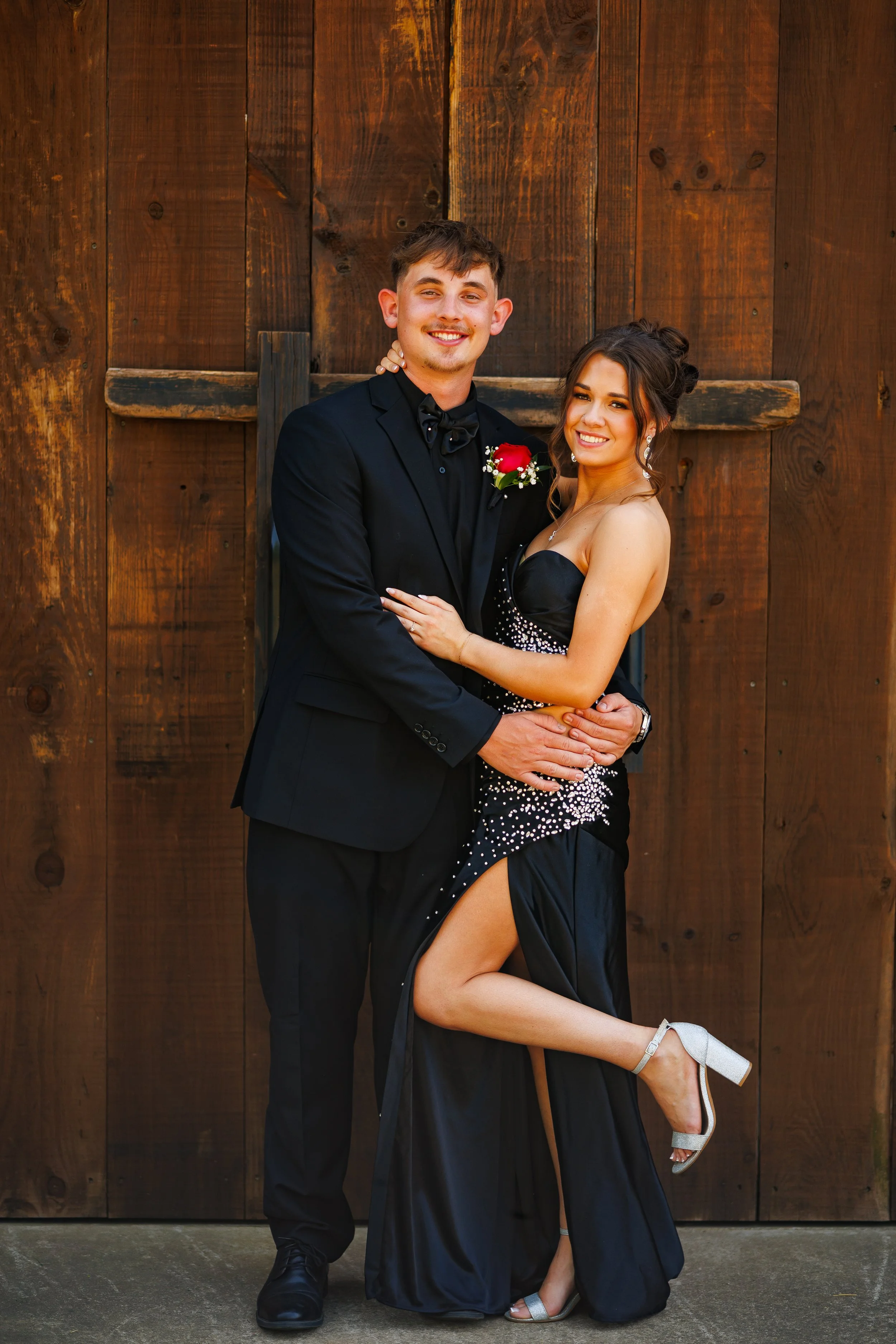 A young couple in formal attire posing in front of a wooden barn door. The man is in a black tuxedo with a boutonniere, and the woman is wearing a black dress with a thigh-high slit and silver high heels.