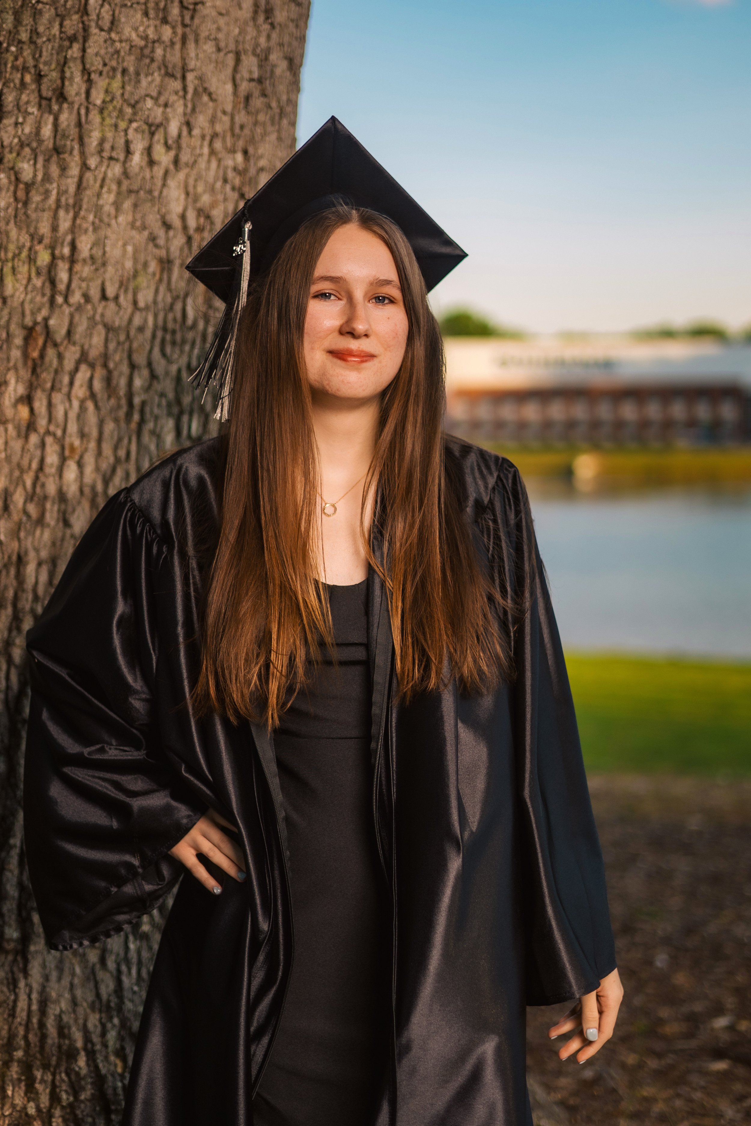 Young woman wearing a black graduation cap and gown standing outdoors near a tree with a lake and buildings in the background during sunset.