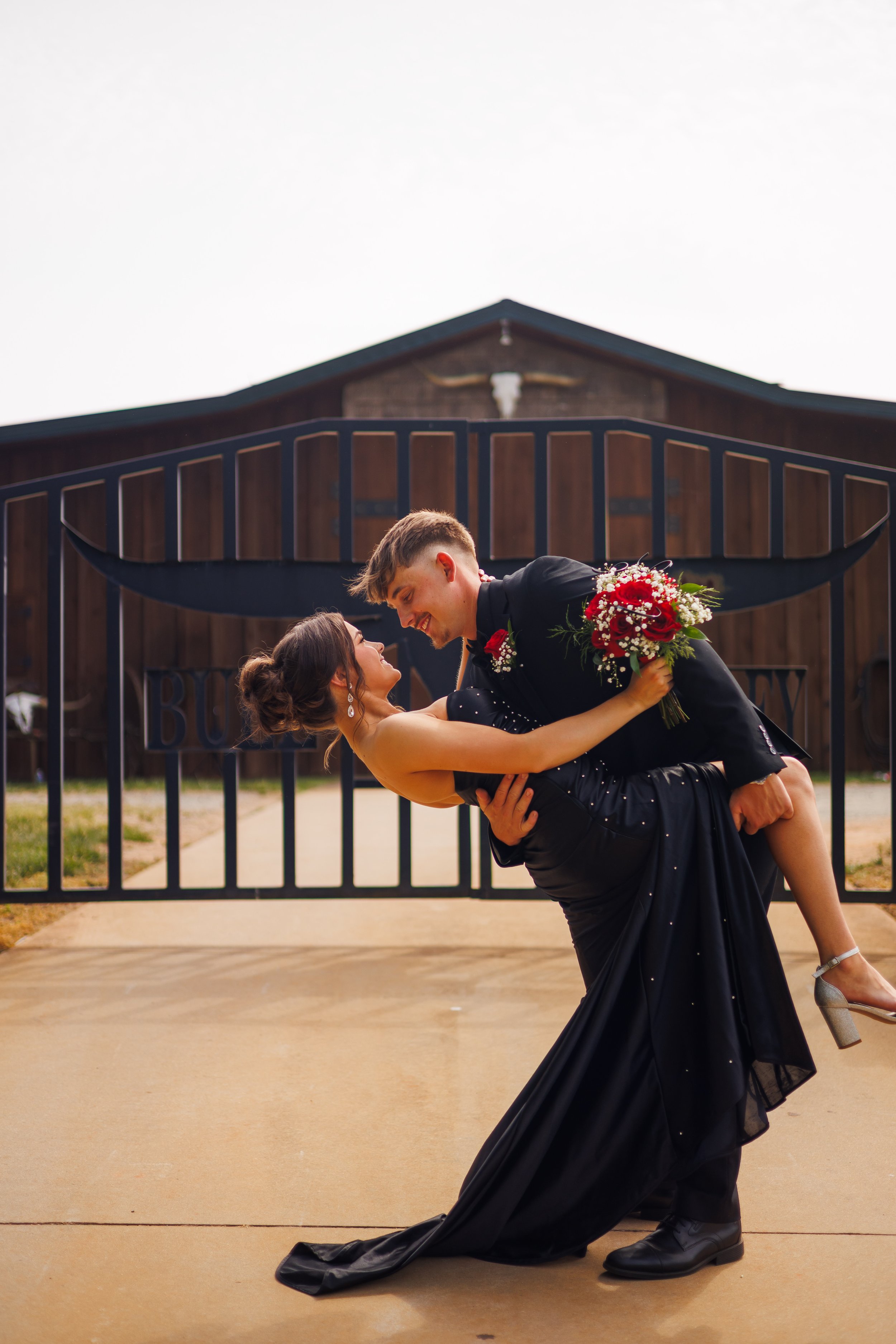 A couple dressed in formal attire sharing a dance outside a barn, with the man lifting the woman in his arms. The woman holds a bouquet of red and white flowers and smiles at the man, while the man leans close, smiling.
