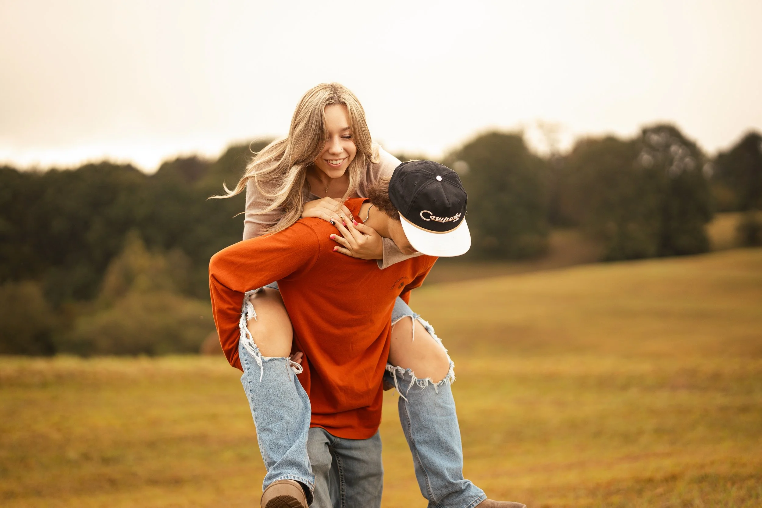 A woman with long blonde hair smiling while giving a piggyback ride to a man wearing a black cap and orange shirt in an outdoor field with trees in the background