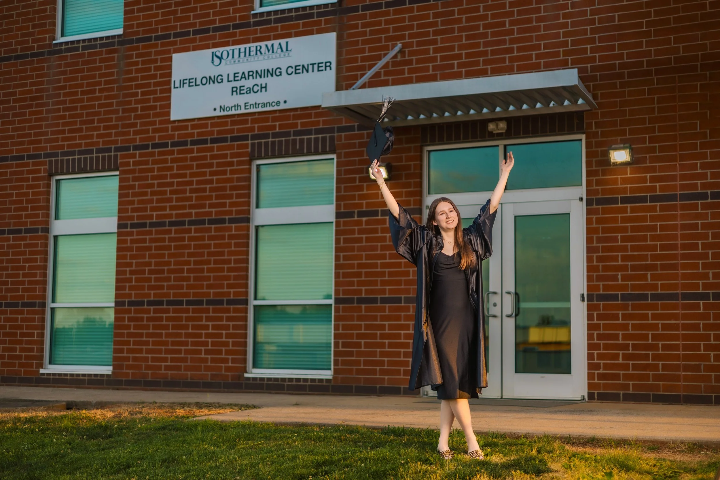 A young woman in a black graduation gown celebrating outside a brick building with a sign that reads 'Isothermal Community College, Lifelong Learning Center ReAch, North Entrance.' She is smiling, holding her graduation cap in the air, and has raised