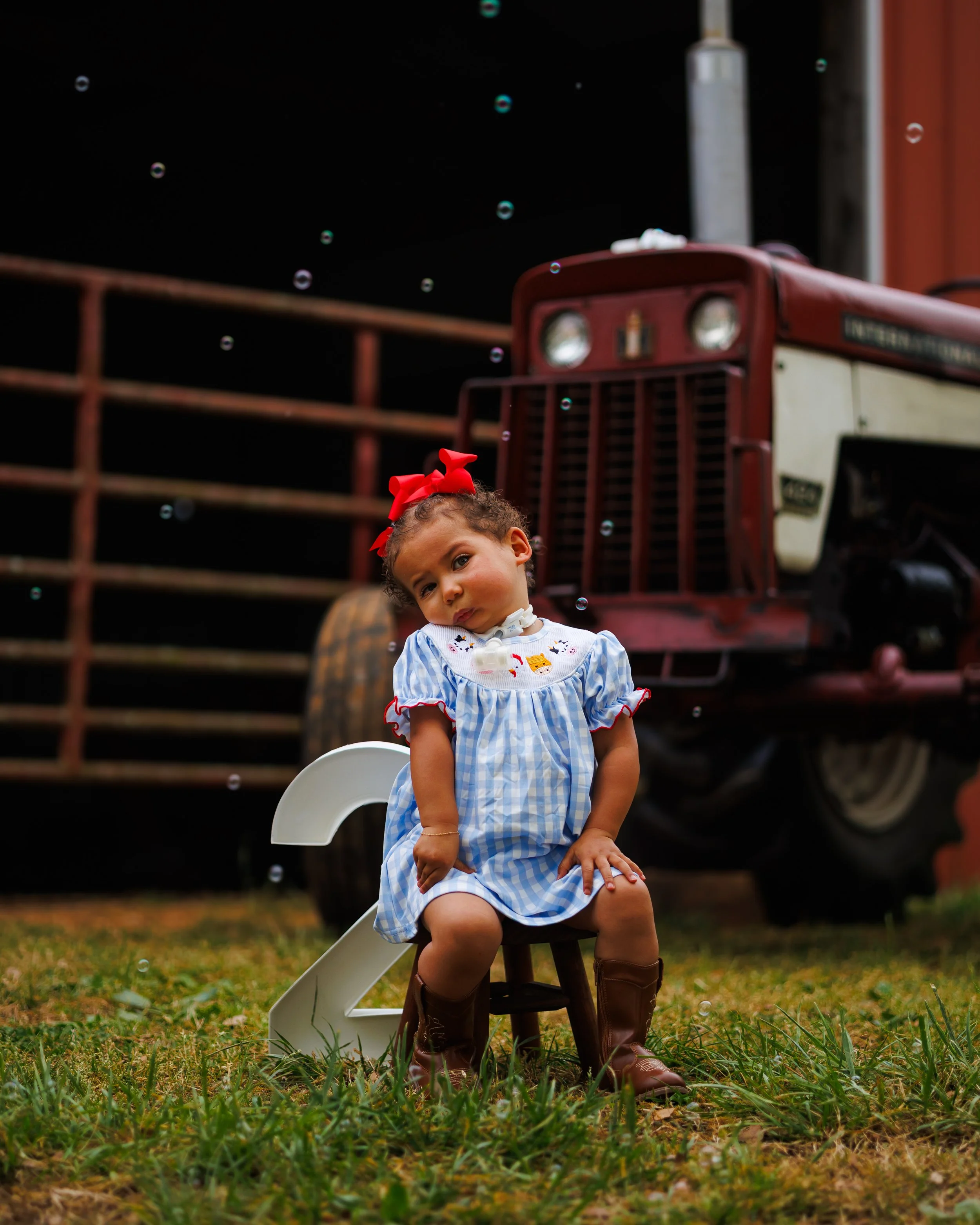 A young girl with curly hair, wearing a blue and white checkered dress, brown boots, and a red bow in her hair, sitting on a wooden chair outdoors in front of a vintage red tractor, with the number 2 and a white plastic chair nearby.