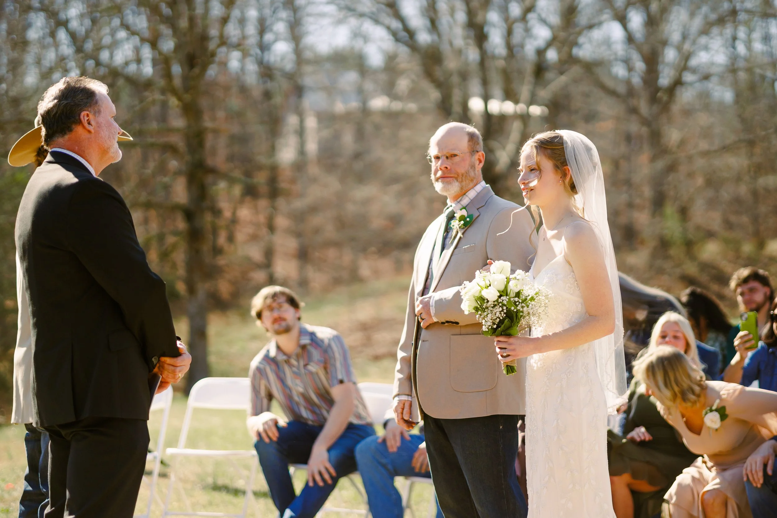 A bride in a white wedding dress holding a bouquet of white flowers stands next to a groom in a gray suit during an outdoor wedding ceremony. An officiant is facing them, holding their hands, with guests seated and watching in the background on a sun