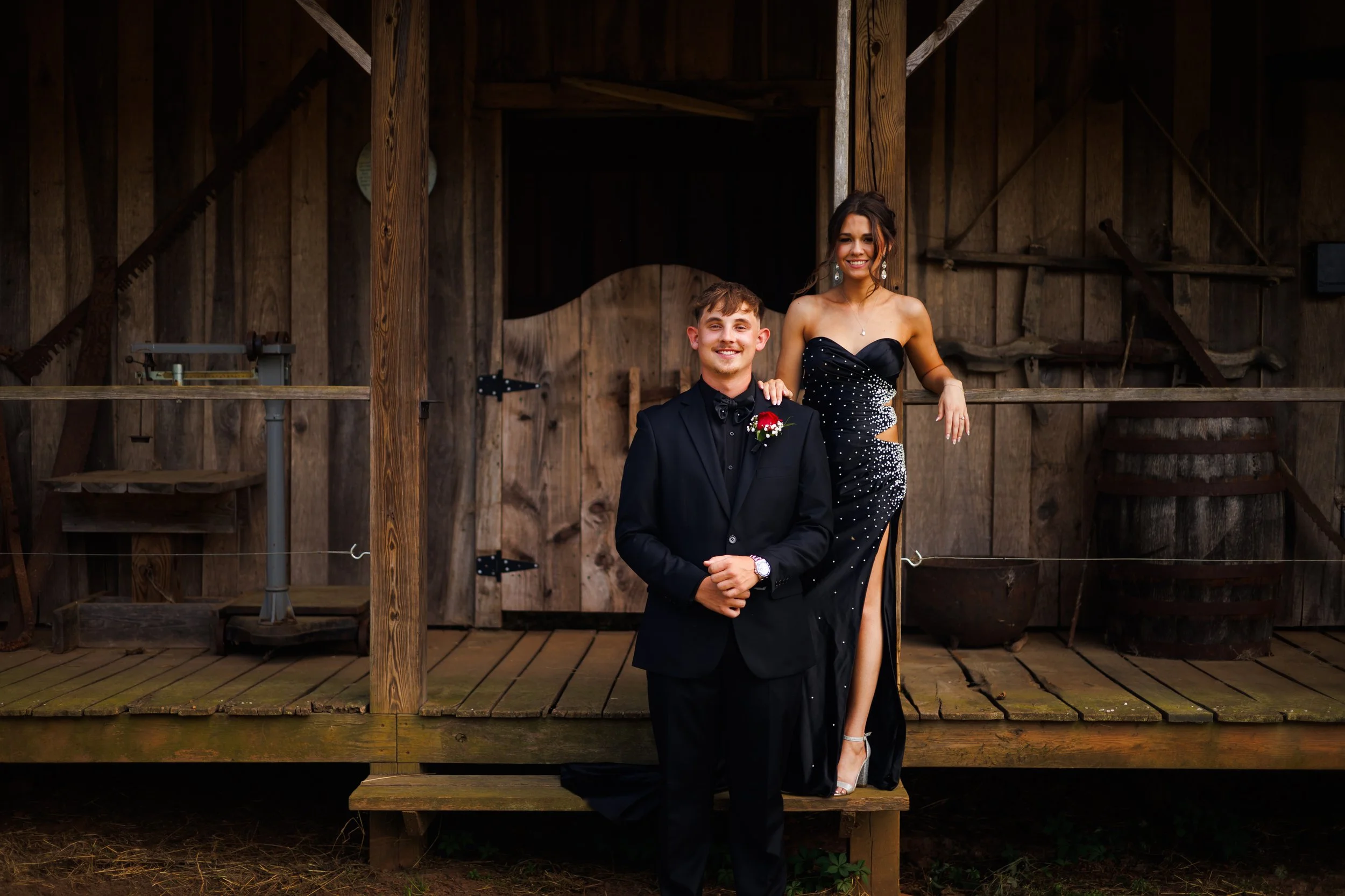 A young man in a black tuxedo with a boutonniere and a young woman in a black and silver gown with jewelry pose together in front of a rustic wooden barn.